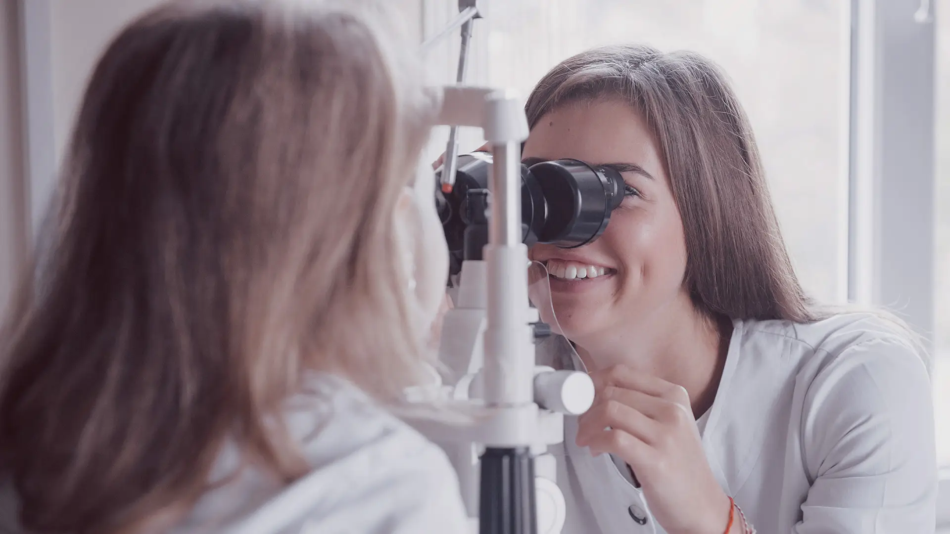 A smiling female optometrist examines a patient’s eyes using a slit lamp in a bright room. The patient, with long light brown hair, sits facing the device whilst the optometrist looks through the microscope, focusing on the patient’s eye.