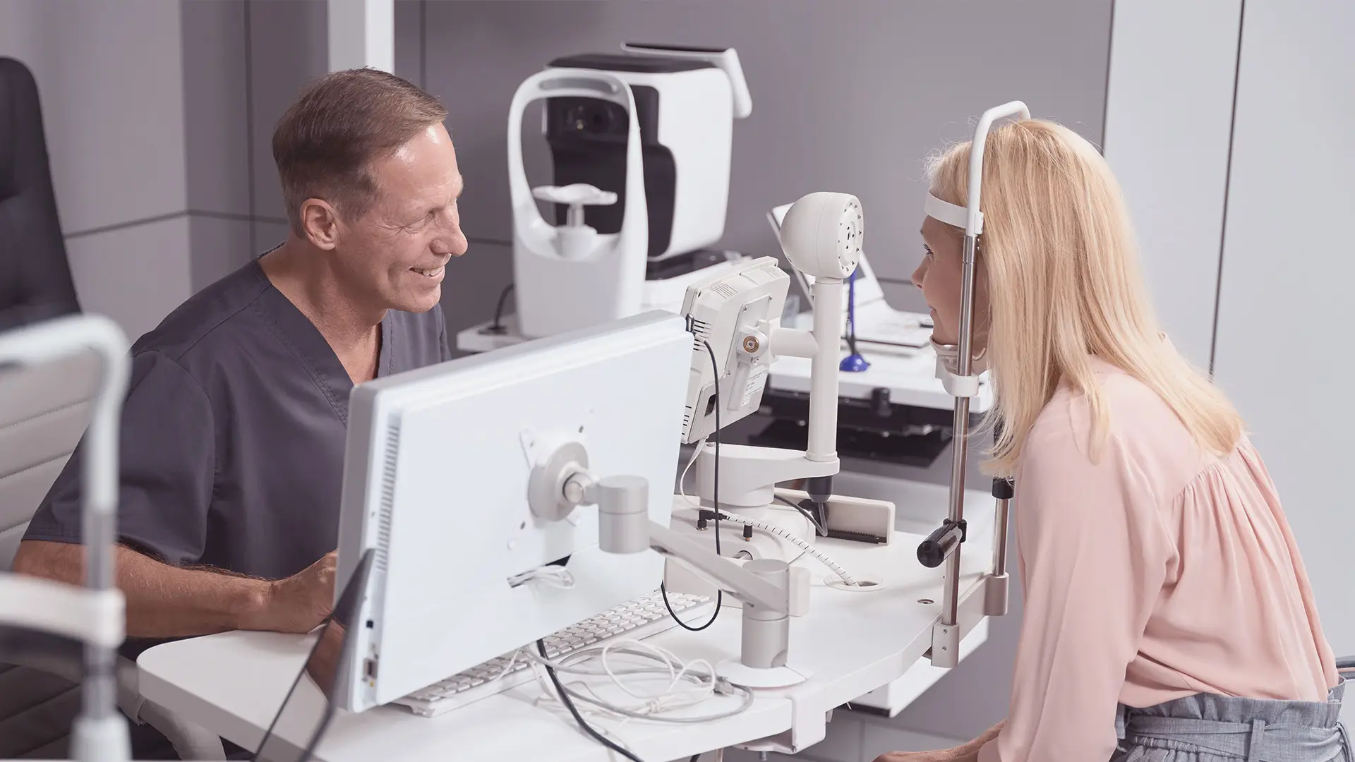 An eye doctor and a female patient sit opposite each other in a clinic. The patient rests her chin on a slit lamp device for an eye examination, while the doctor smiles and observes her. Medical equipment and a computer are on the desk between them.