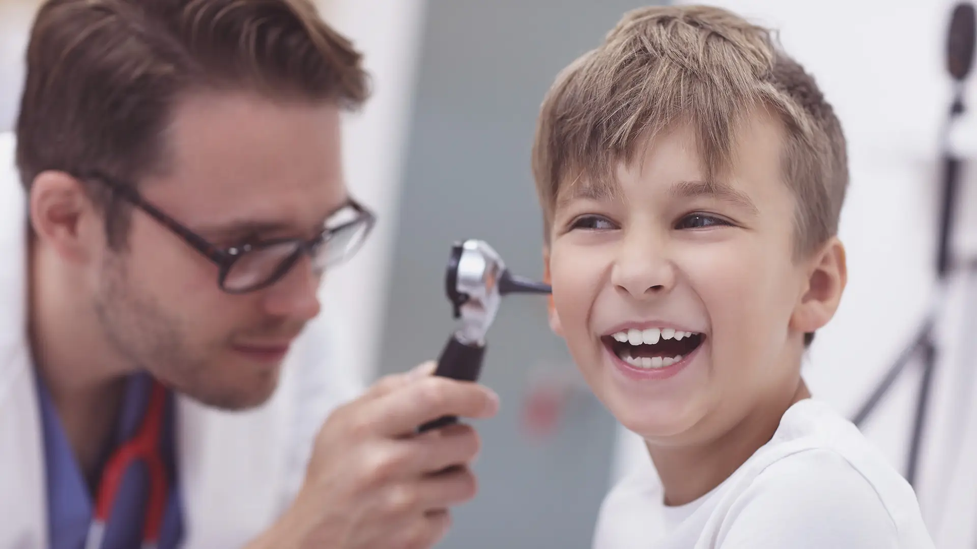 A smiling boy sits while a doctor examines his ear with an otoscope. The doctor wears glasses and a white coat. The boy looks happy and relaxed in a brightly lit medical surgery. The background is blurred, focusing on their interaction.