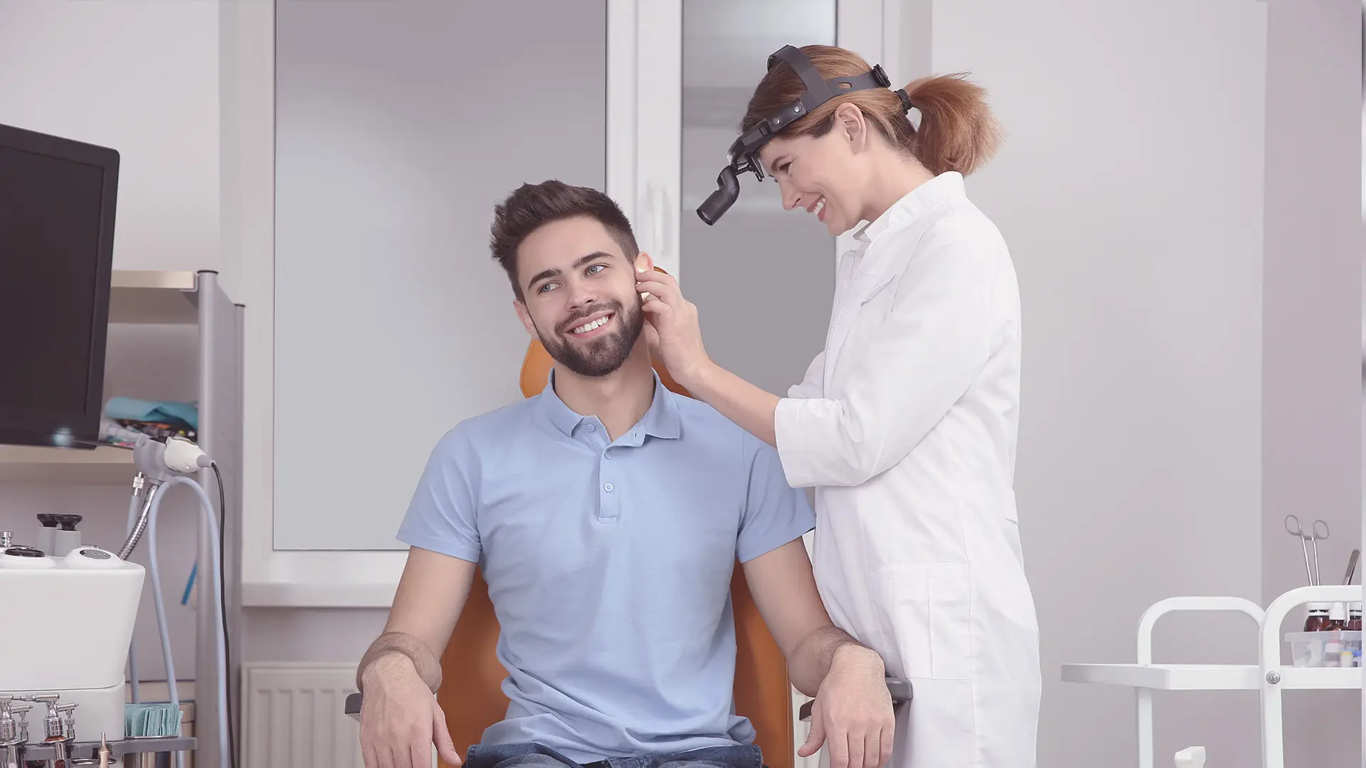 A smiling male patient sits in a clinic chair whilst a female doctor in a white coat and head torch examines his ear. Medical equipment is visible around them, and the setting is a modern, bright examination room. Both appear relaxed and comfortable.