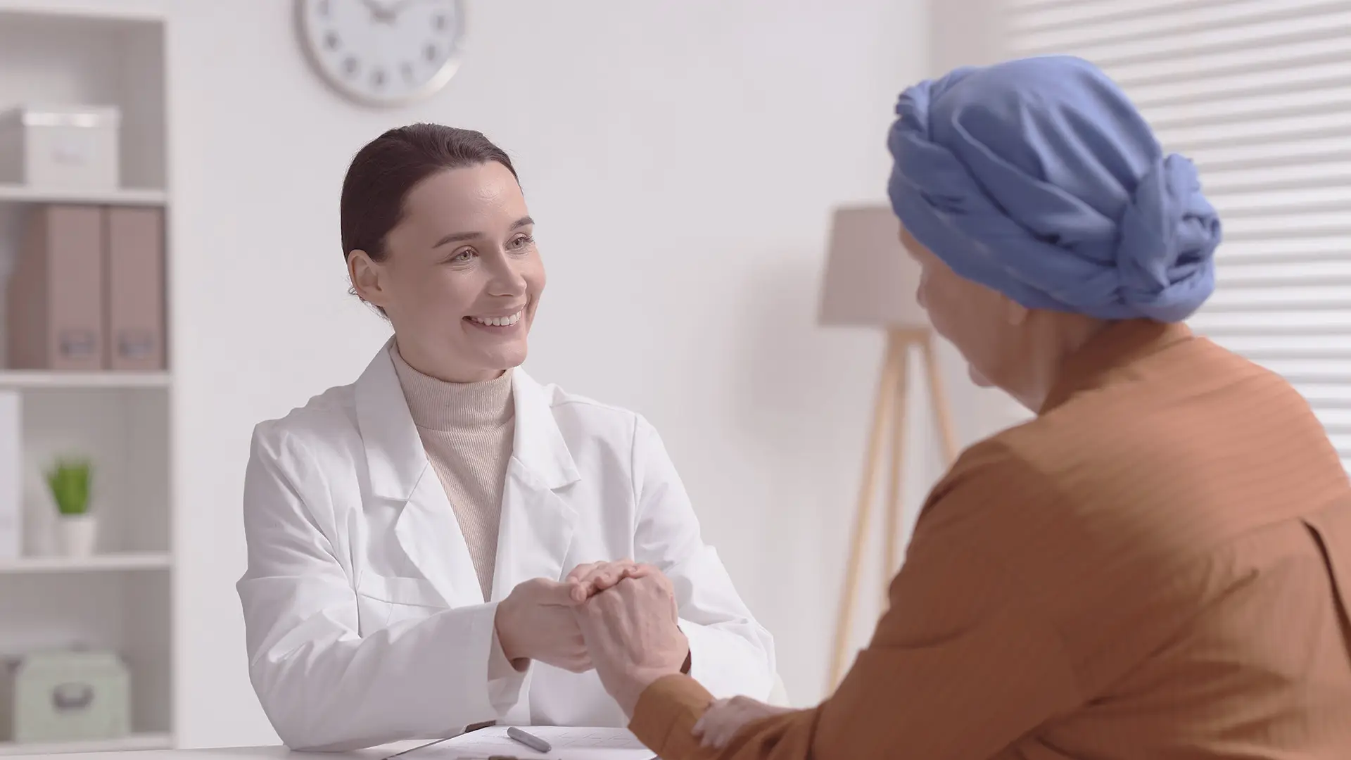 A female doctor in a white coat warmly holds the hands of a patient wearing a blue headscarf and brown top. They are seated across a desk in a bright office, suggesting a supportive medical consultation. A clock and shelves are visible in the background.