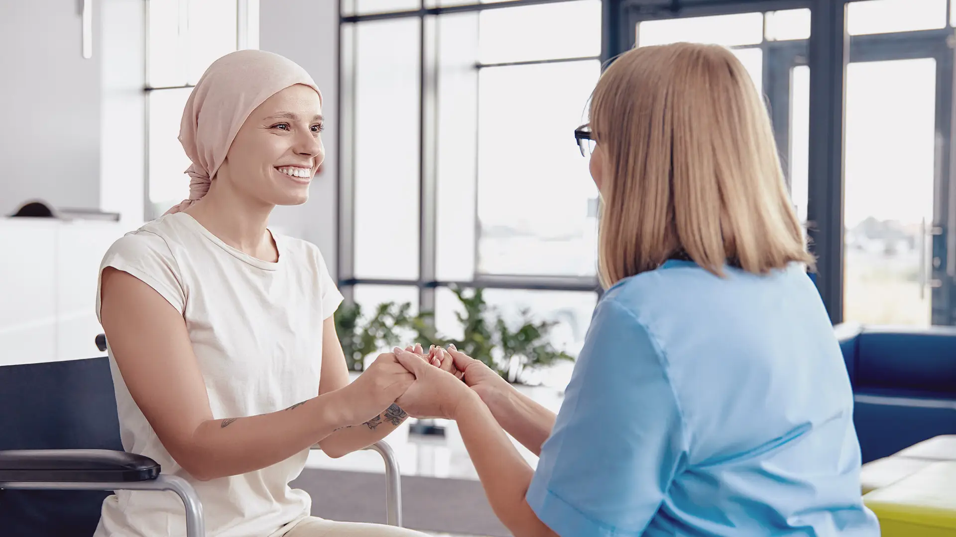 A smiling woman wearing a headscarf sits in a wheelchair, holding hands with a healthcare professional in blue scrubs. They face each other in a bright room with large windows and plants, suggesting support and encouragement.