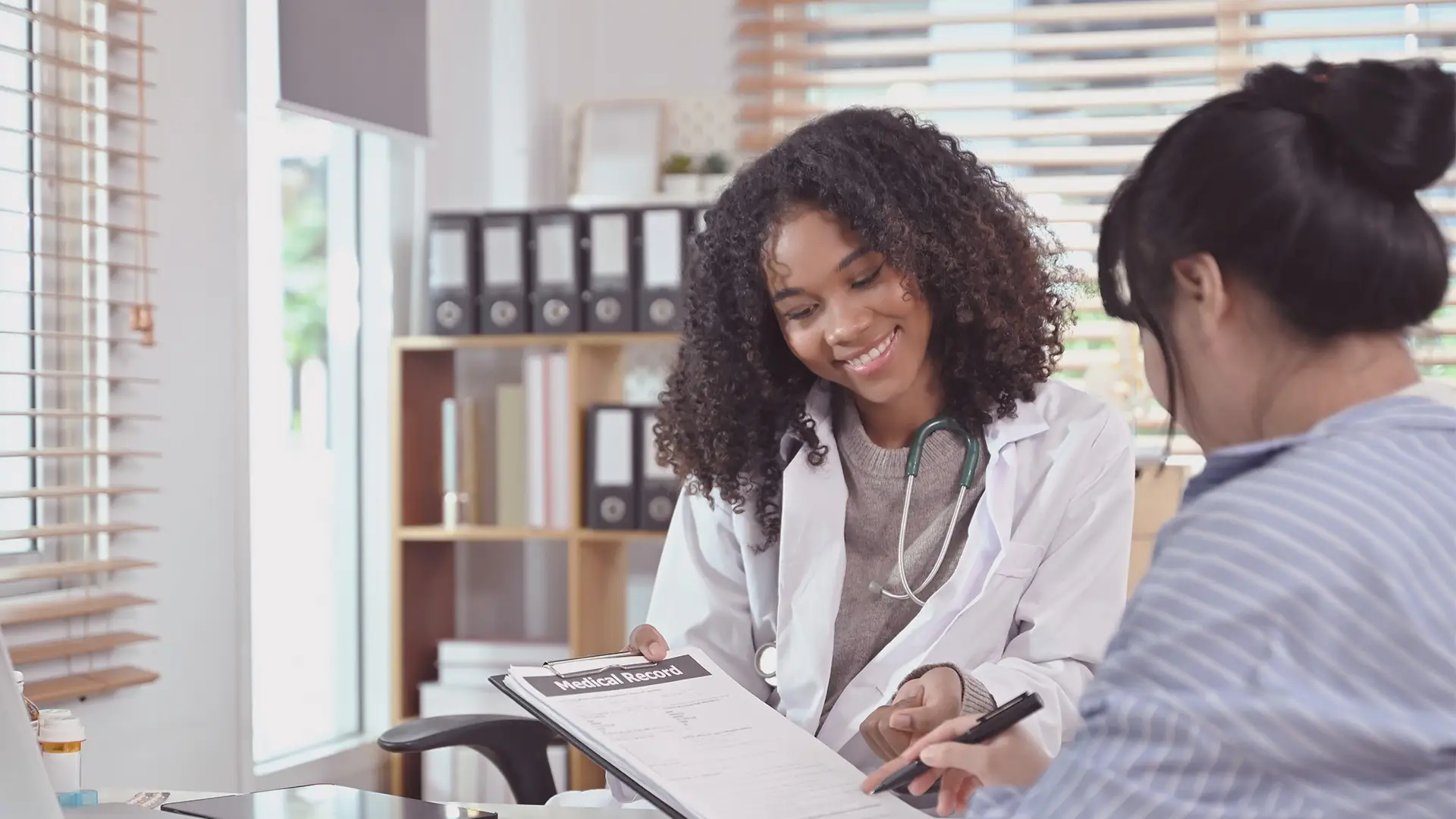 A smiling doctor with curly hair wearing a white coat and stethoscope hands a clipboard labelled “Medical Record” to a patient in striped clothing, who is holding a pen, in a bright office with shelves and blinds in the background.