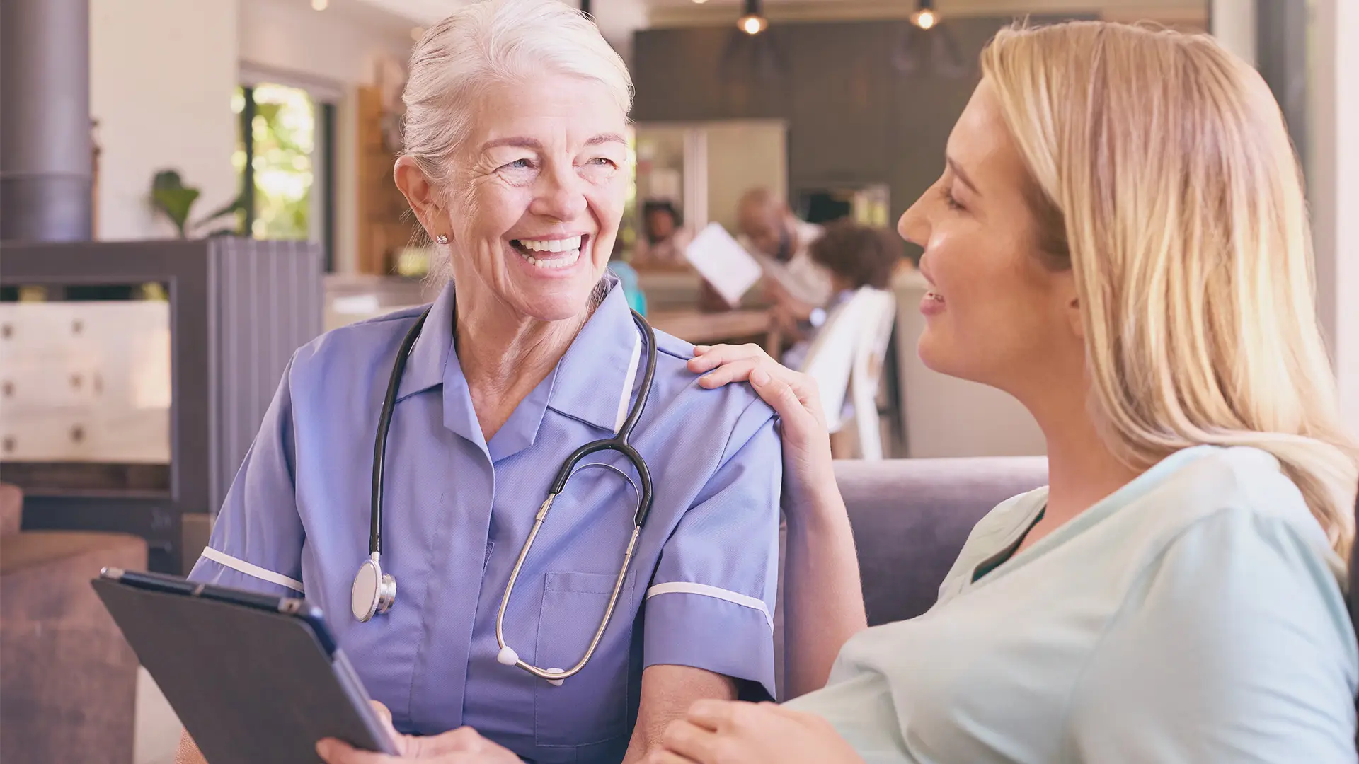 An older female nurse with short grey hair, wearing a blue uniform and stethoscope, smiles warmly whilst holding a clipboard and talking to a younger woman with blonde hair, who gently touches the nurse’s shoulder. They sit in a bright, cosy room.