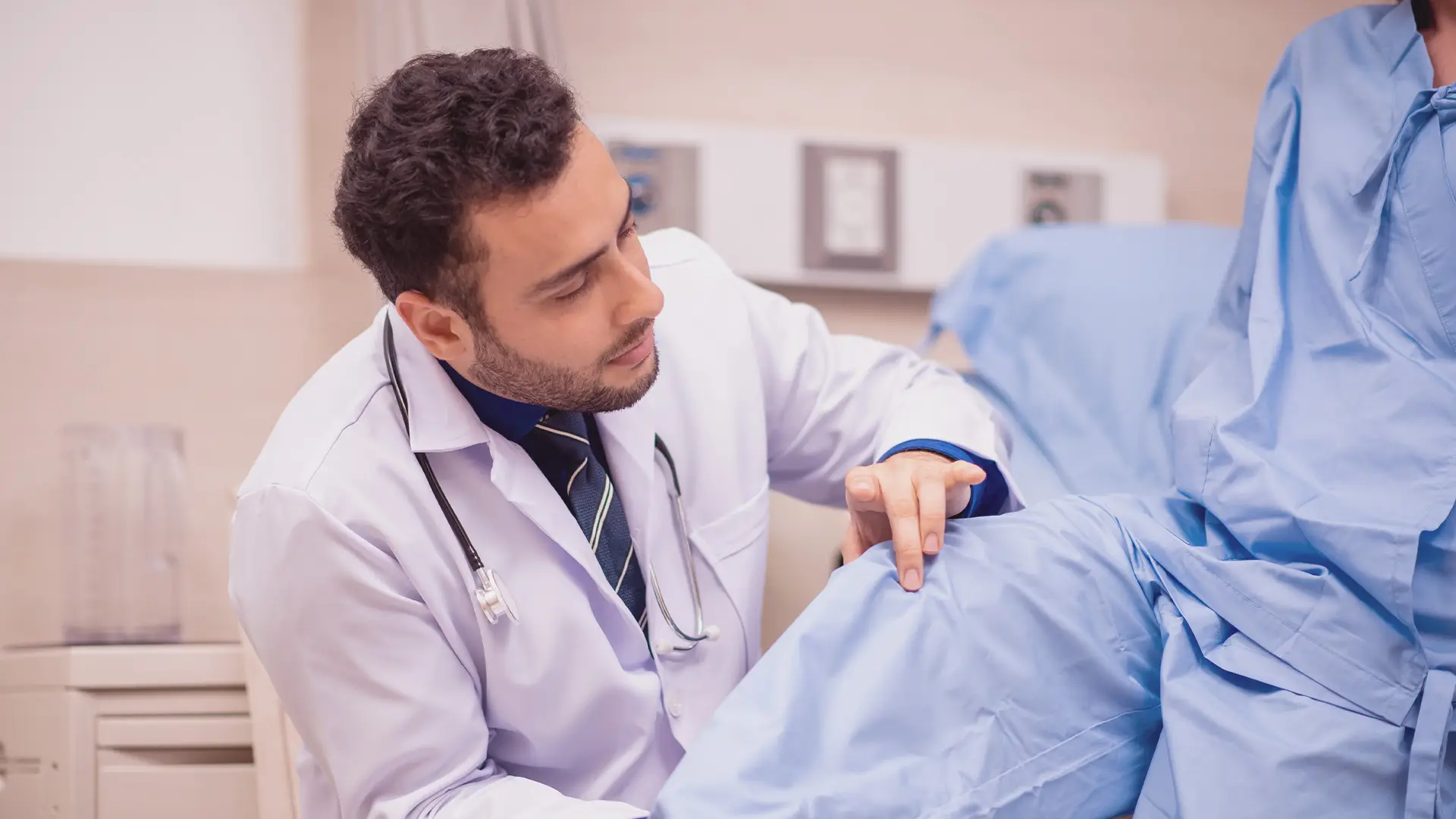 A male doctor in a white coat examines a patient’s knee in a medical setting. The doctor is focused, gently touching the patient’s covered leg while the patient sits on an examination bed wearing a blue gown. Medical equipment is visible in the background.