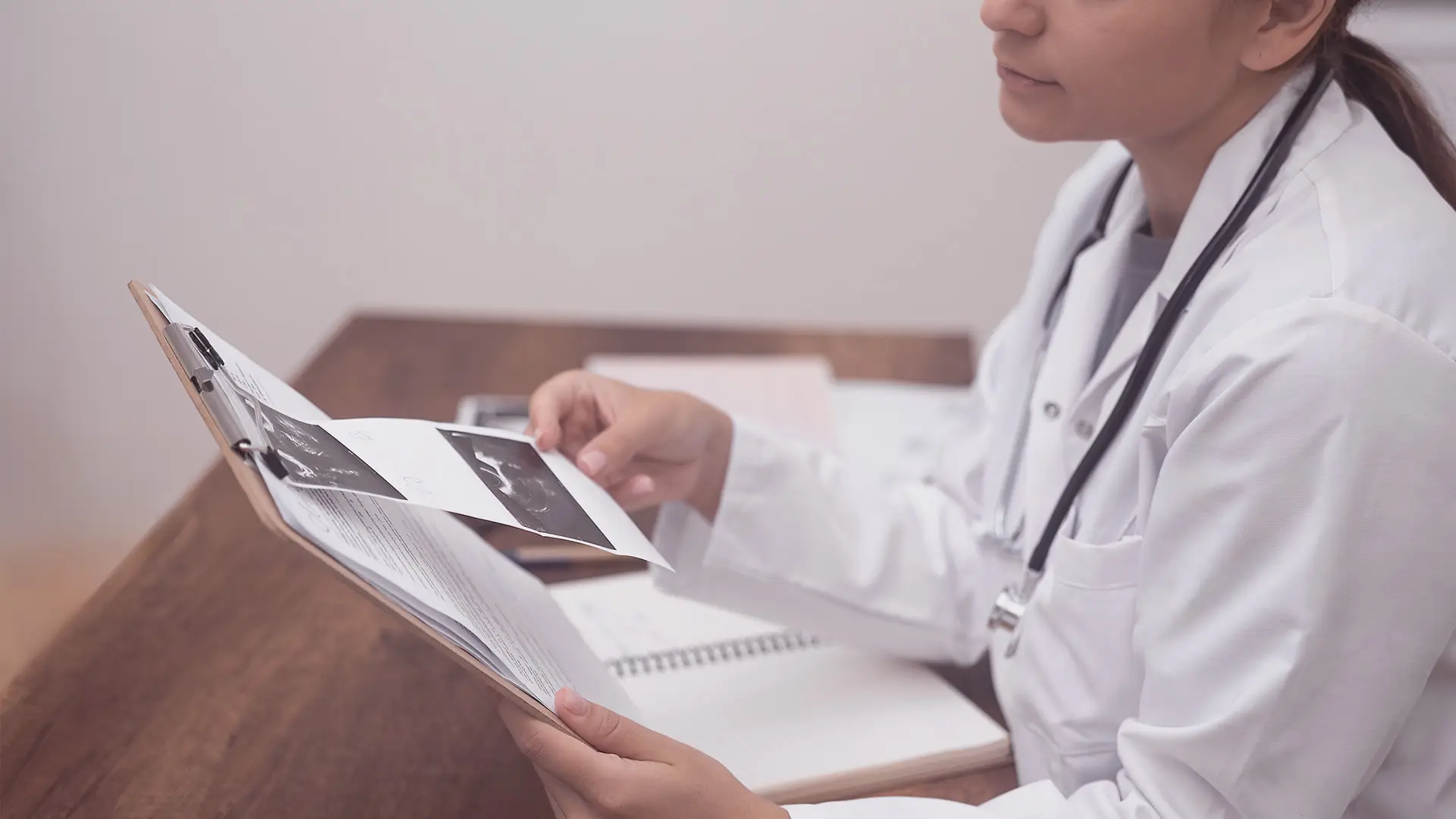 A doctor in a white coat sits at a desk holding a clipboard and examining ultrasound images. The desk has an open notebook and some papers. The doctor is partially visible from the side, with a stethoscope round their neck.