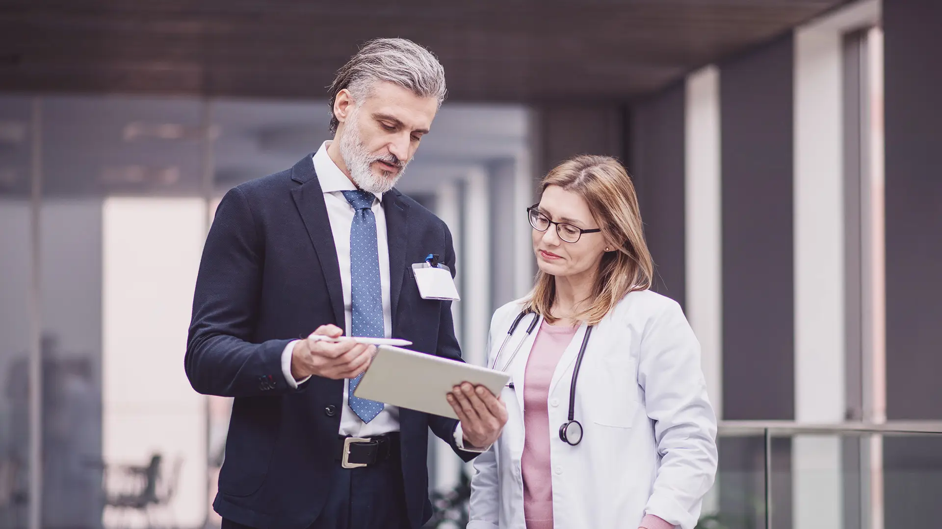 A man in a suit holds a tablet and discusses its contents with a woman in a white coat and stethoscope. They stand indoors, appearing focused and engaged in conversation, possibly collaborating in a professional or healthcare setting.