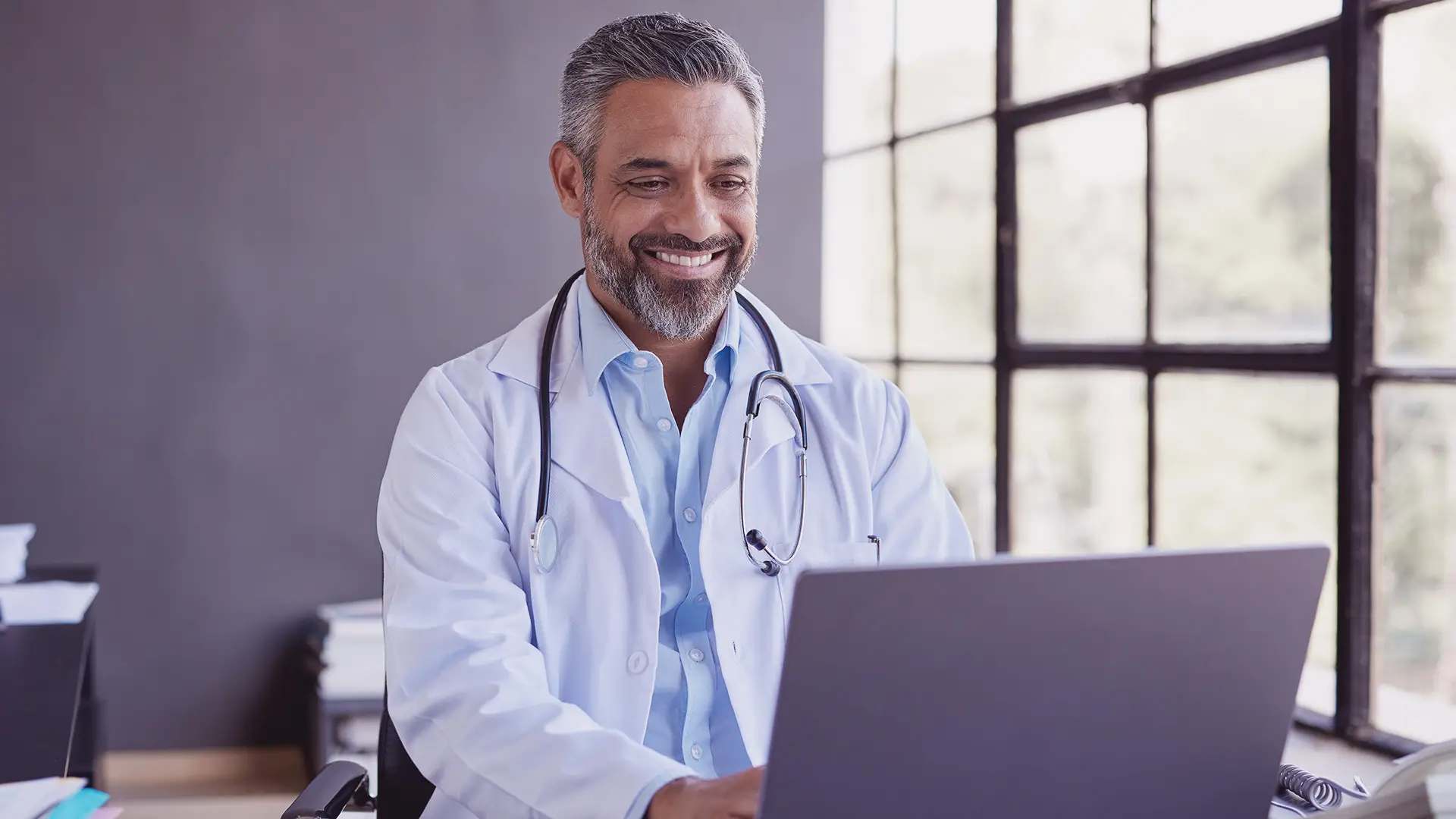 A smiling male doctor with grey hair and beard, wearing a white lab coat and stethoscope, sits at a desk using a laptop. Large windows behind him let in natural light, and he appears focused and approachable in a modern office setting.