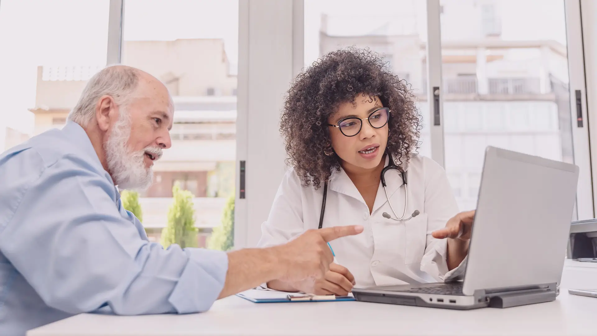 A female doctor with curly hair and glasses sits at a desk, showing information on a laptop to an older male patient with a beard. Both are engaged in discussion, gesturing towards the screen, with large windows and buildings in the background.