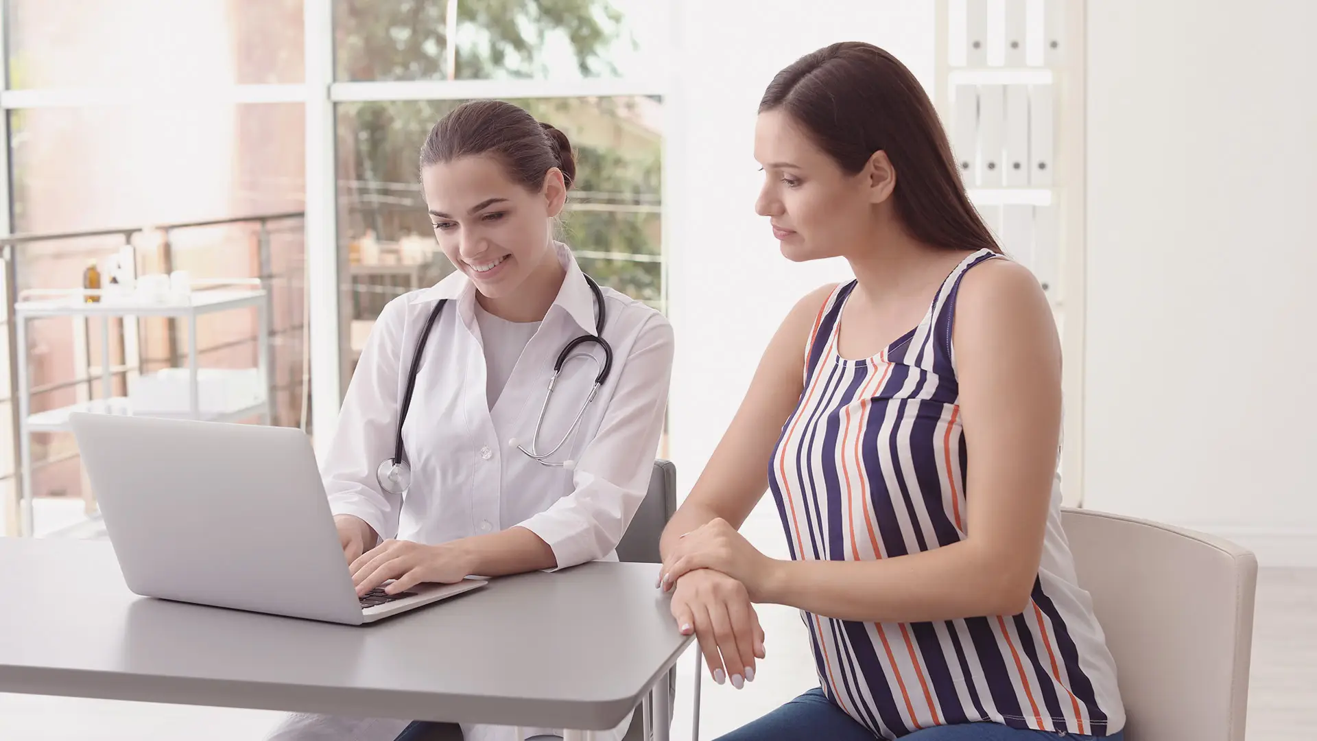 A female doctor in a white coat and stethoscope sits at a desk with a laptop, smiling whilst showing something on the screen to a seated woman in a striped sleeveless top. They are in a bright medical surgery with shelves and large windows.
