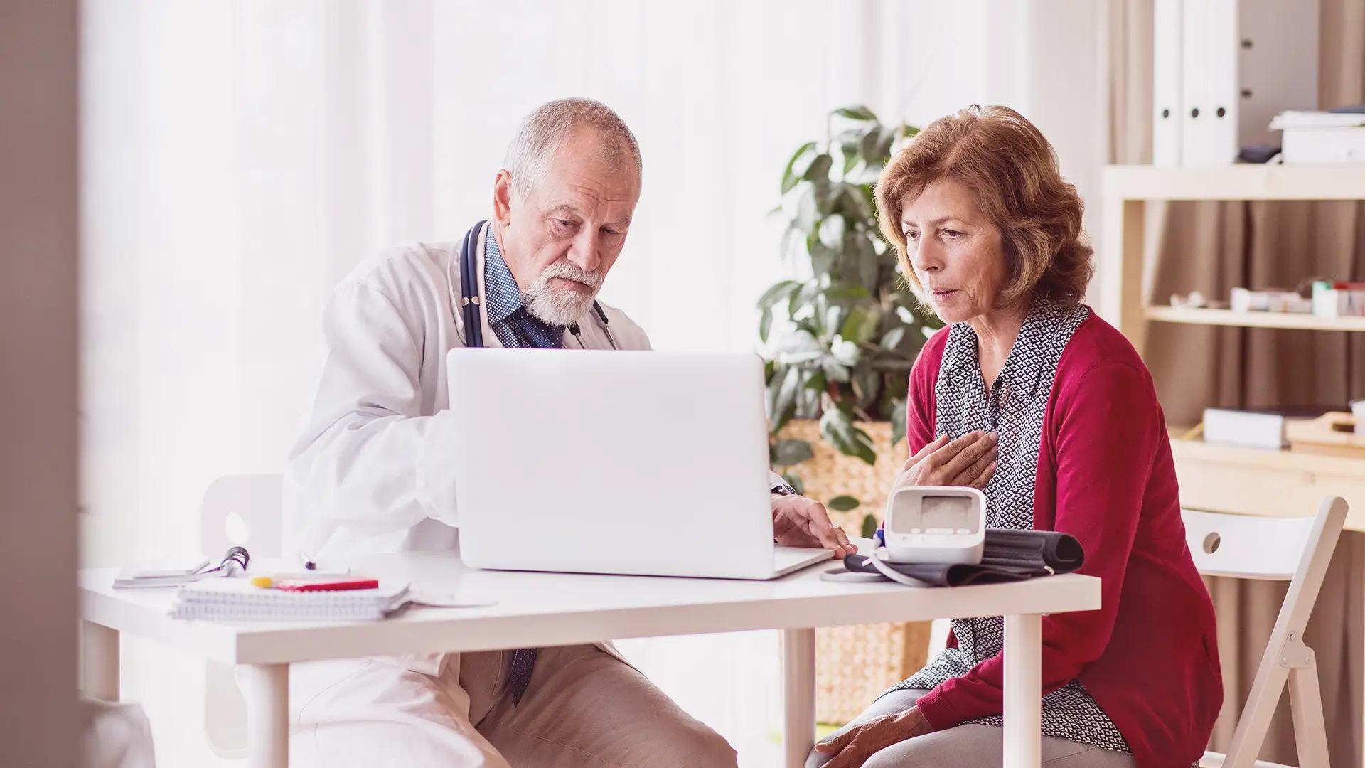 An older male doctor shows information on a laptop to an older woman, who looks concerned and touches her chest. They sit at a white desk with a blood pressure monitor and notepad in a bright, modern medical surgery with plants and shelves.