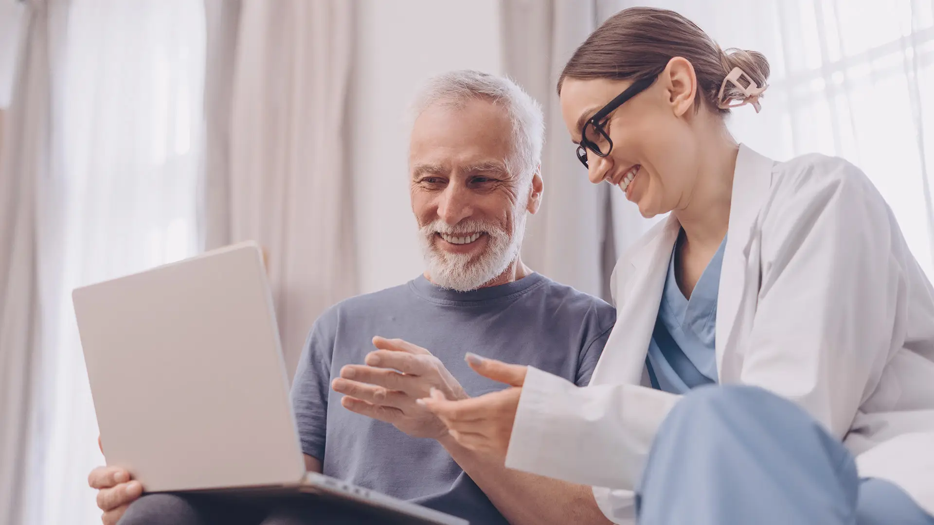 An older man with grey hair and beard smiles while sitting next to a female doctor in glasses and a white coat. They look at a laptop together, appearing happy and engaged, in a bright room with light curtains.