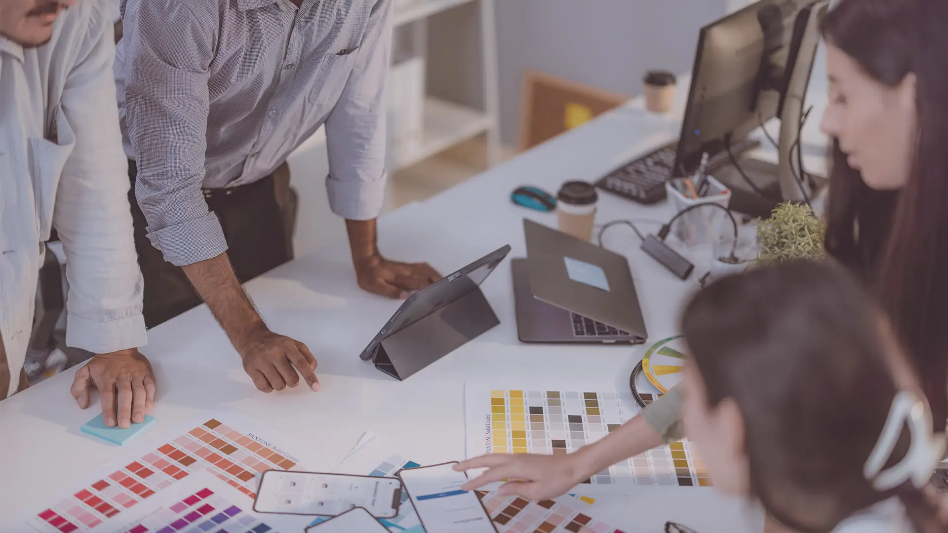 Four people gather around a white desk covered with colour swatches, charts, a mobile phone, a tablet, and a laptop. One person points at the swatches while others look on, suggesting a collaborative design or planning meeting in a modern office space.