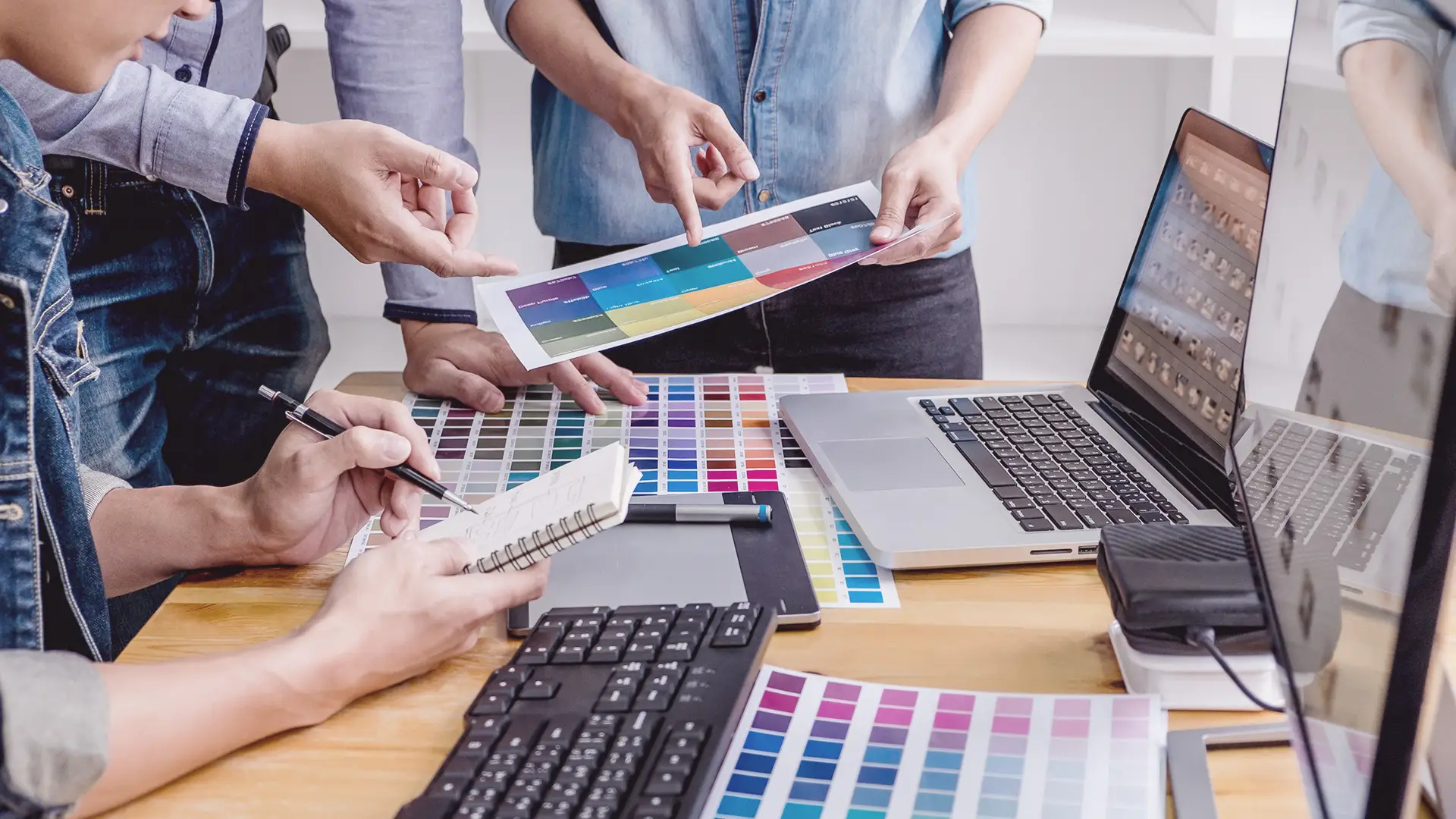 Three people collaborate at a desk covered with colour swatches, laptops, and a keyboard. One person writes in a notepad, another points at a colour chart, and the third holds a paper, suggesting a design or creative planning discussion.