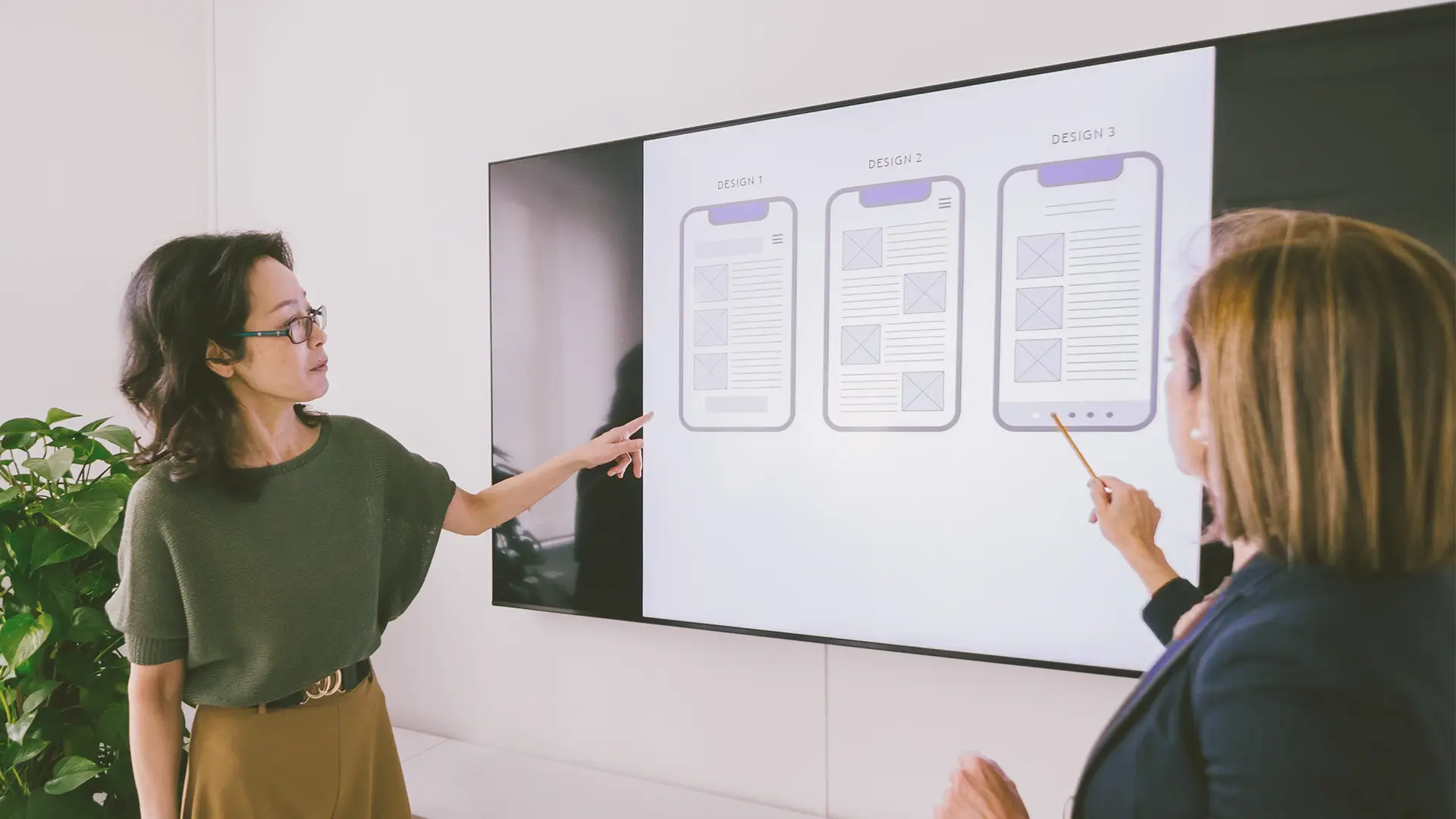 Two women in business attire stand in front of a large screen displaying three smartphone app wireframes labelled “DESIGN 1,” “DESIGN 2,” and “DESIGN 3.” Each woman points at a different design, appearing to discuss the options in an office setting.