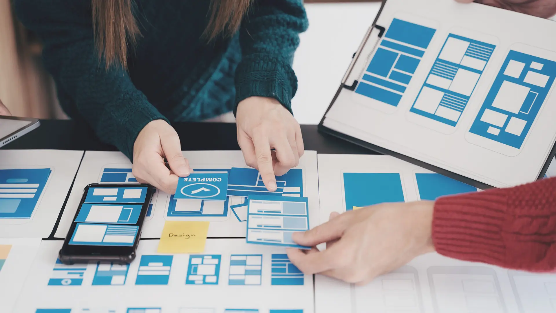 Three people collaborate at a table covered with blue and white website or app wireframes and mock-ups, pointing to layouts and discussing design. One person holds a clipboard with more layout options, emphasising teamwork in user interface design.