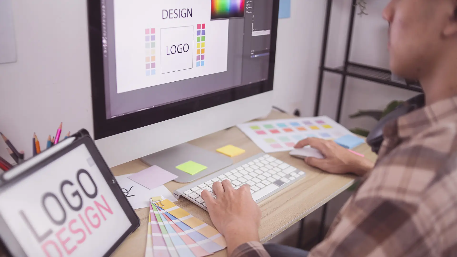 A person uses a desktop computer to design a logo, shown on the screen with various colour palettes. A colour swatch fan, sticky notes, and a tablet displaying LOGO DESIGN are on the desk. The person holds a keyboard and mouse.