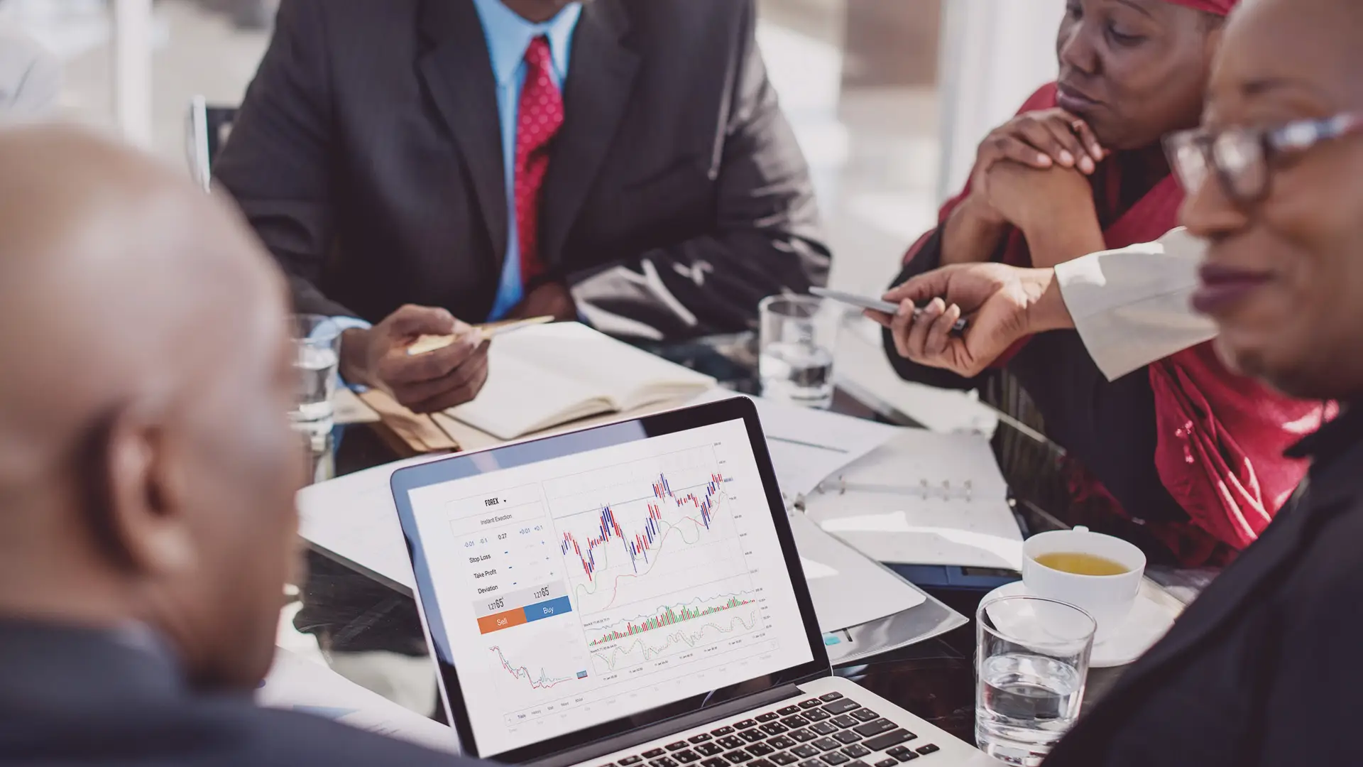 Four people in business attire sit around a glass table in discussion. A laptop displaying a colourful line graph with charts is in the foreground. Notebooks, papers, glasses of water, and a cup of tea are also on the table, indicating a work meeting.