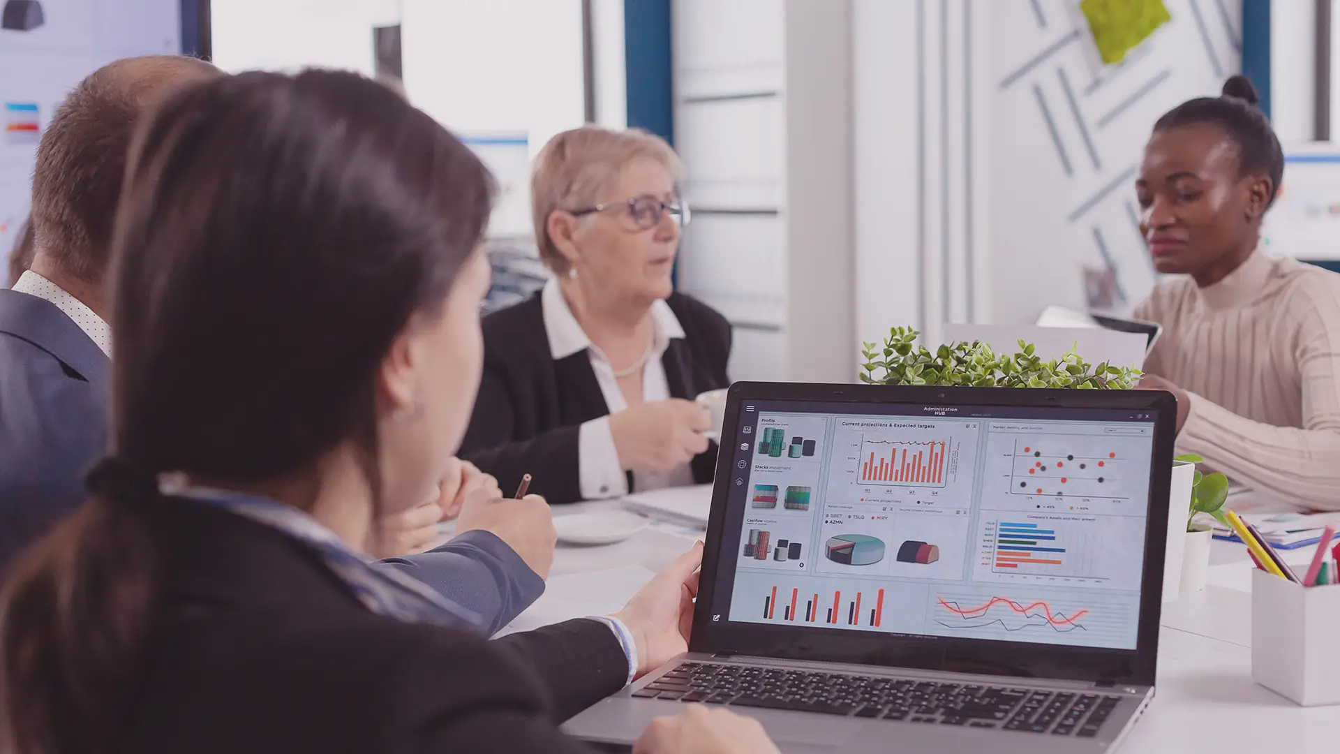 A group of four people sit at a table in an office, discussing. In the foreground, a laptop screen displays various colourful financial charts and graphs. One person takes notes, whilst others listen and talk, with papers and plants on the table.
