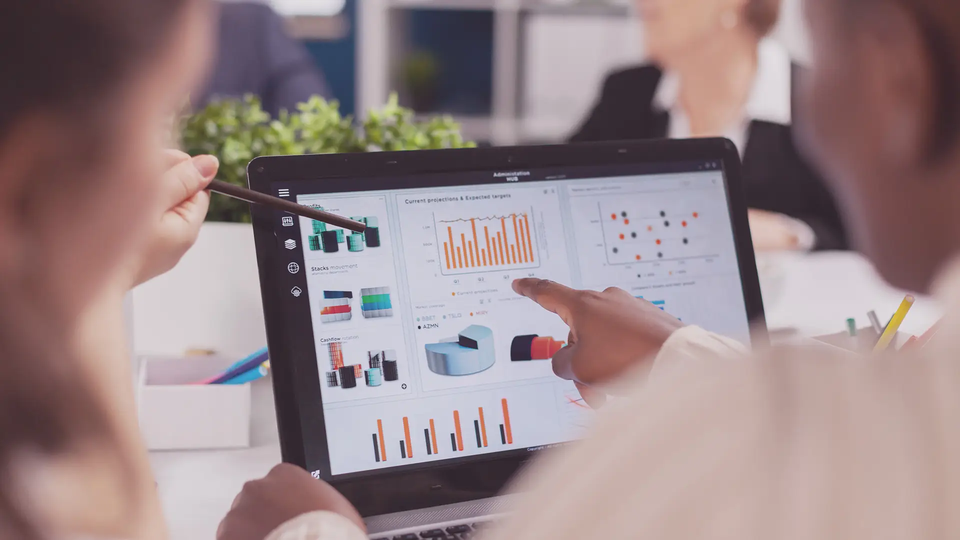 Two people review data graphs on a laptop screen, including bar charts, pie charts, and line graphs. One person points at the display whilst the other uses a pen to indicate data. A plant and office supplies are visible in the background.