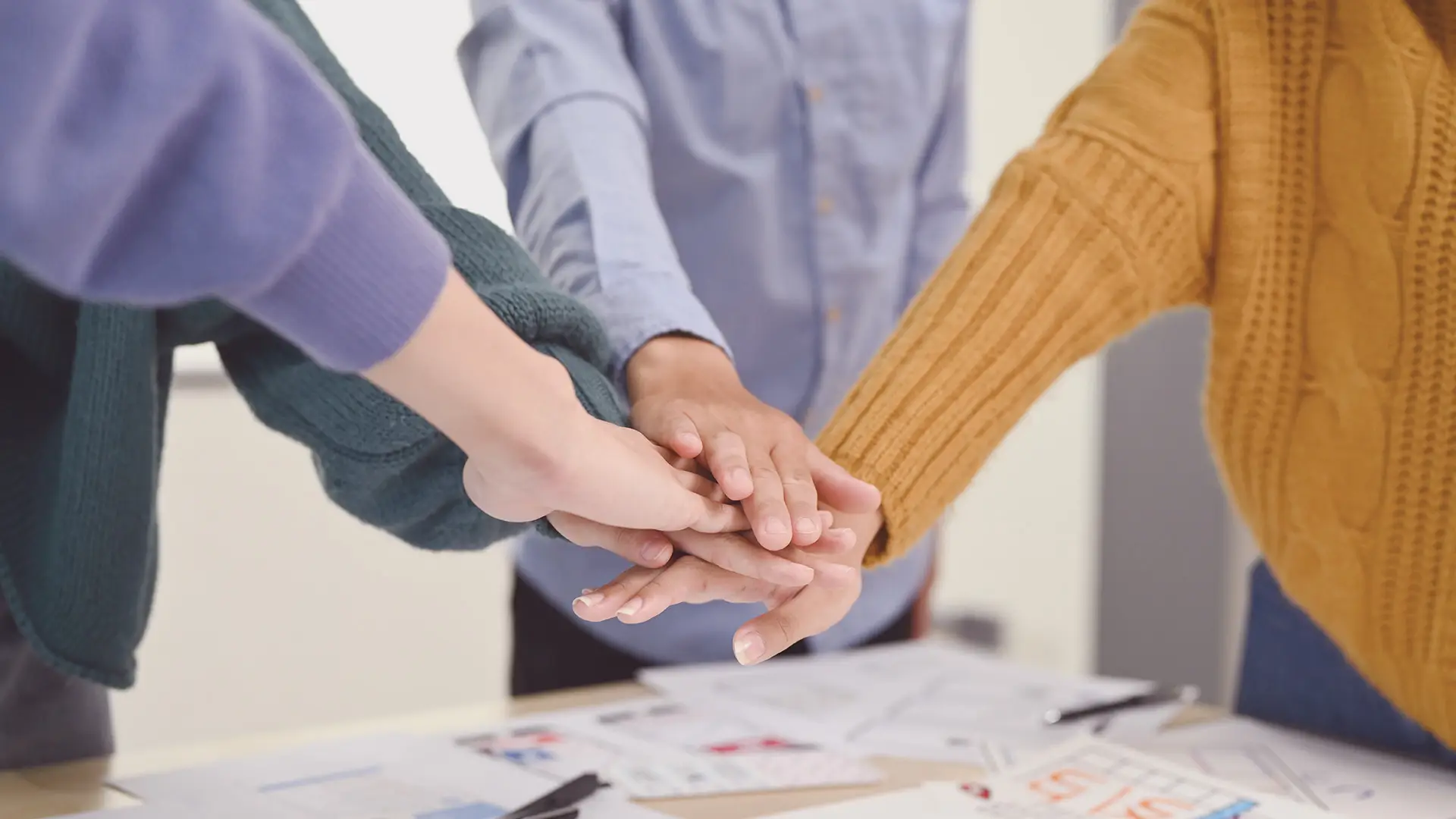 Four people stand around a table, stacking their hands together in a gesture of teamwork. They wear casual clothes: jumpers in green and yellow, and a blue button-up shirt. Papers and pens are scattered on the table beneath their hands.