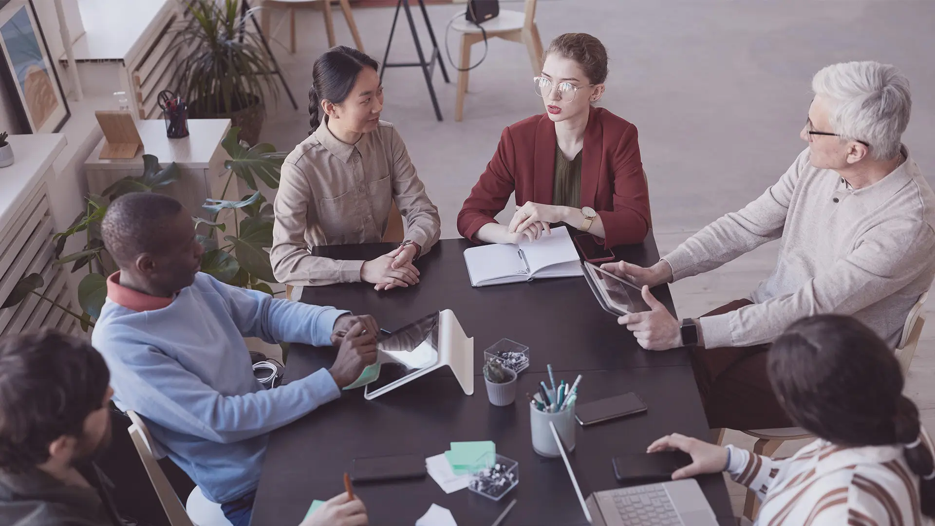 Six people of diverse backgrounds sit around a table in an office. They discuss, take notes, and use digital devices. The group includes men and women, plants decorate the space, and notebooks, tablets, and papers are spread on the table.