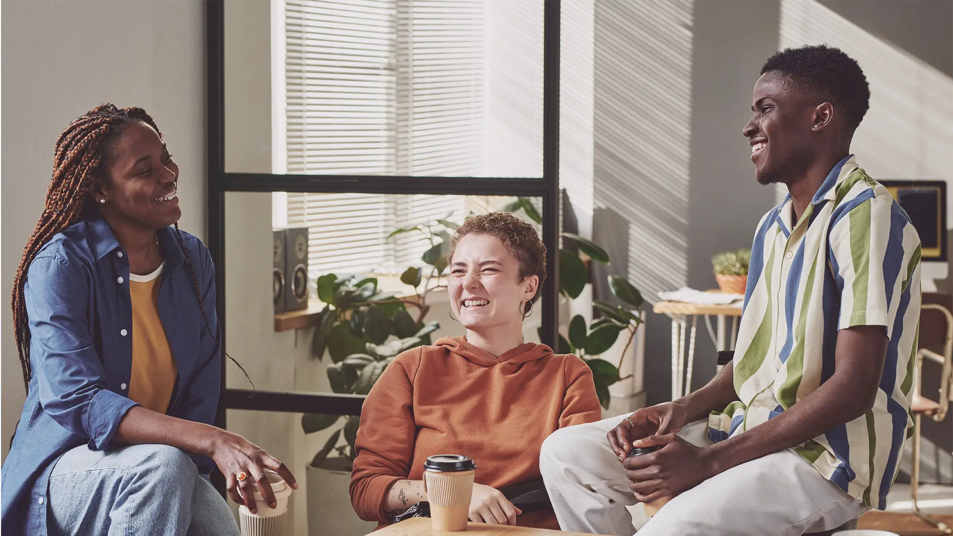 Three people sit together indoors, smiling and talking. One wears a blue shirt and jeans, another wears an orange hoodie, and the third wears a striped shirt. Sunlight shines in through blinds, and green plants decorate the room. A coffee cup sits on the table.