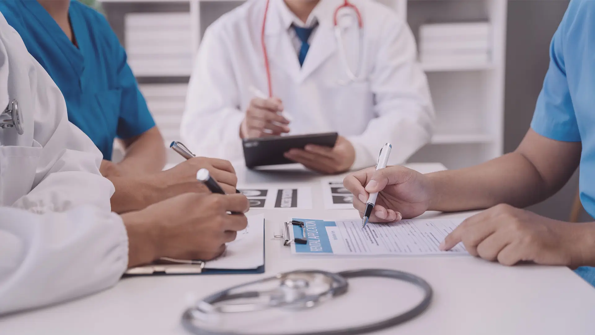 Four healthcare professionals in scrubs and lab coats sit at a table, writing on clipboards and discussing documents. A stethoscope lies on the table, and one person holds a tablet. The atmosphere is collaborative and focused.