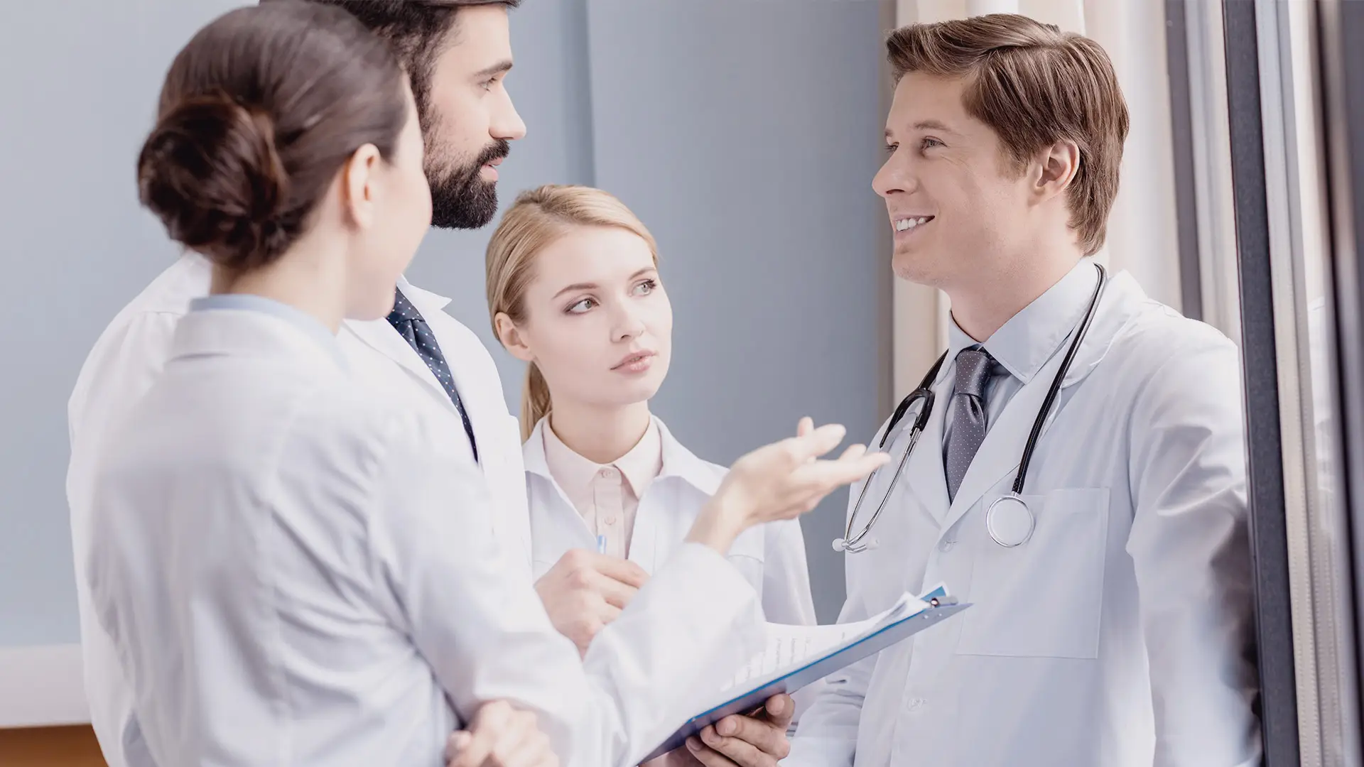 Four doctors in white coats stand together indoors, having a discussion. One doctor holds a clipboard and gestures as they talk. Another doctor, wearing a stethoscope, smiles. The group appears engaged in a professional conversation near a window.