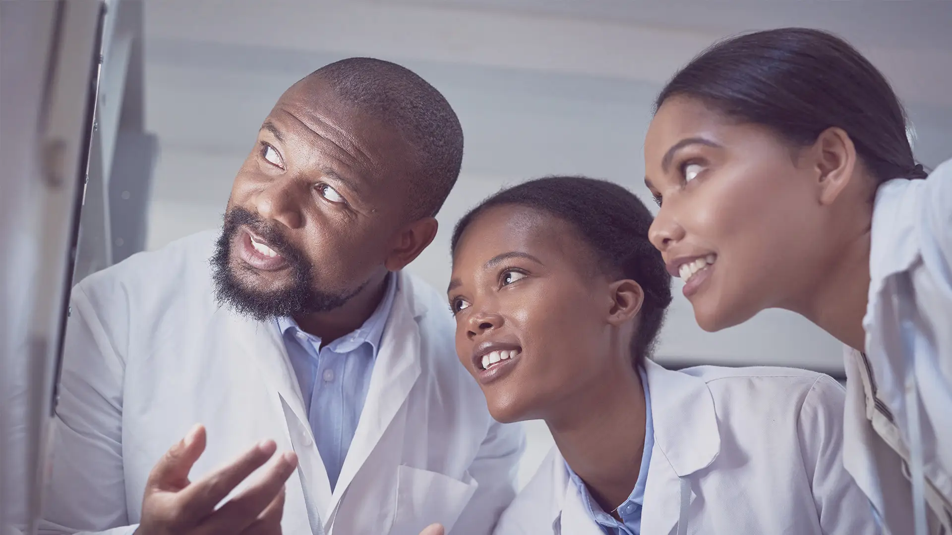 Three people wearing white lab coats closely examine something off-screen. They appear engaged and are smiling. The background is blurred, focusing attention on their attentive expressions and collaborative interaction, suggesting a scientific or medical setting.