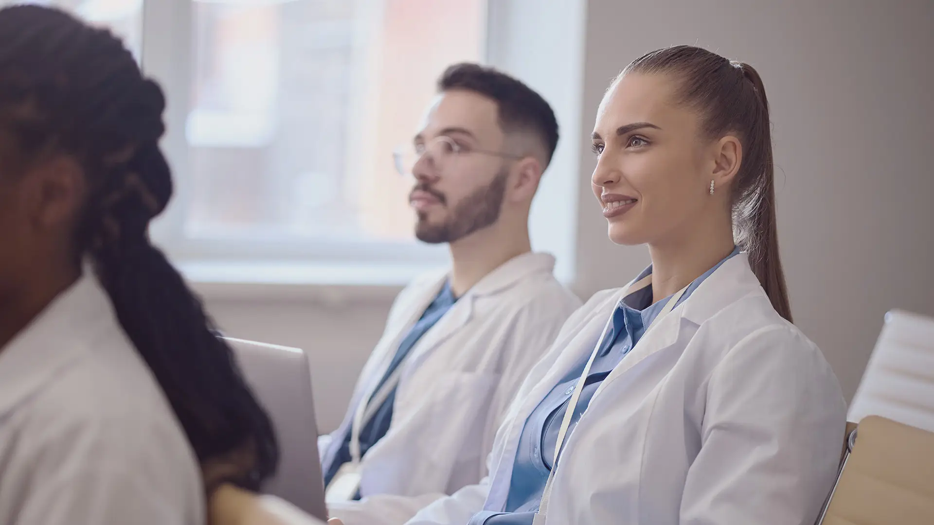 Several young adults in white lab coats sit attentively in a bright classroom. The focus is on a woman with a ponytail smiling, and a man with glasses beside her. Both appear engaged, suggesting a medical or healthcare educational setting.