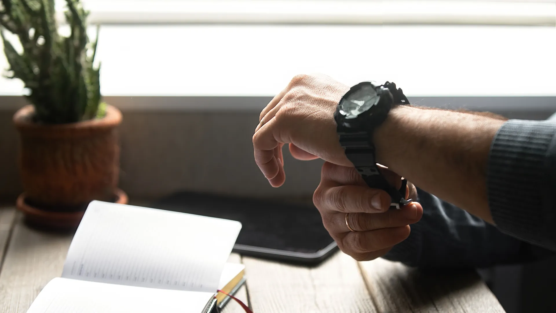 A man wearing a black wristwatch checks the time beside an open notebook and a plant on a desk.