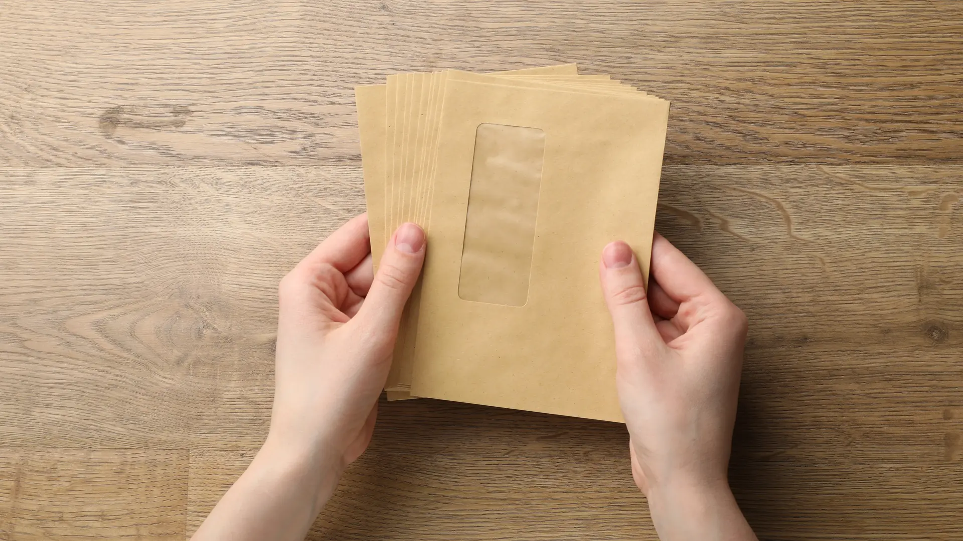 A woman's hands hold a stack of brown window envelopes over a wooden desk.