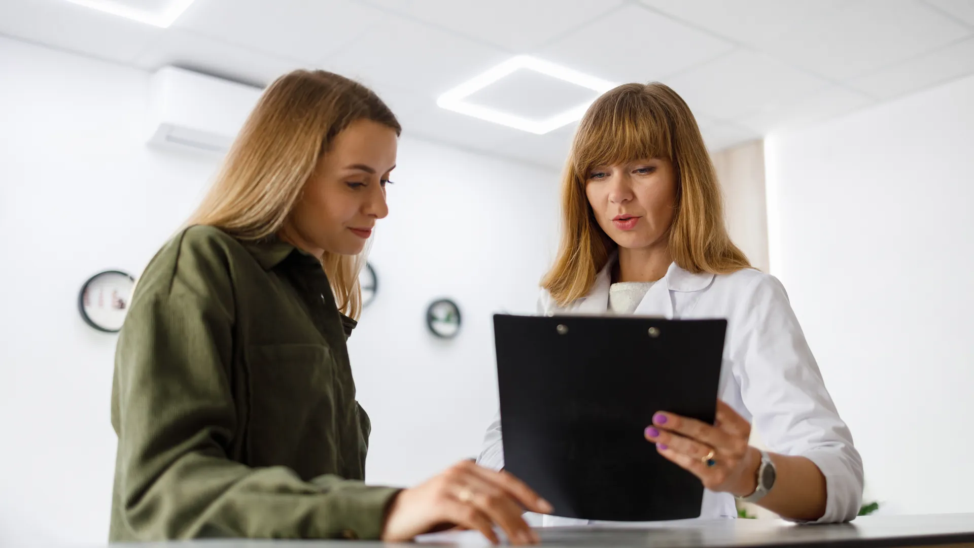 A woman in a green jacket and a doctor in a white coat look together at a form on a black clipboard at a clinic desk.