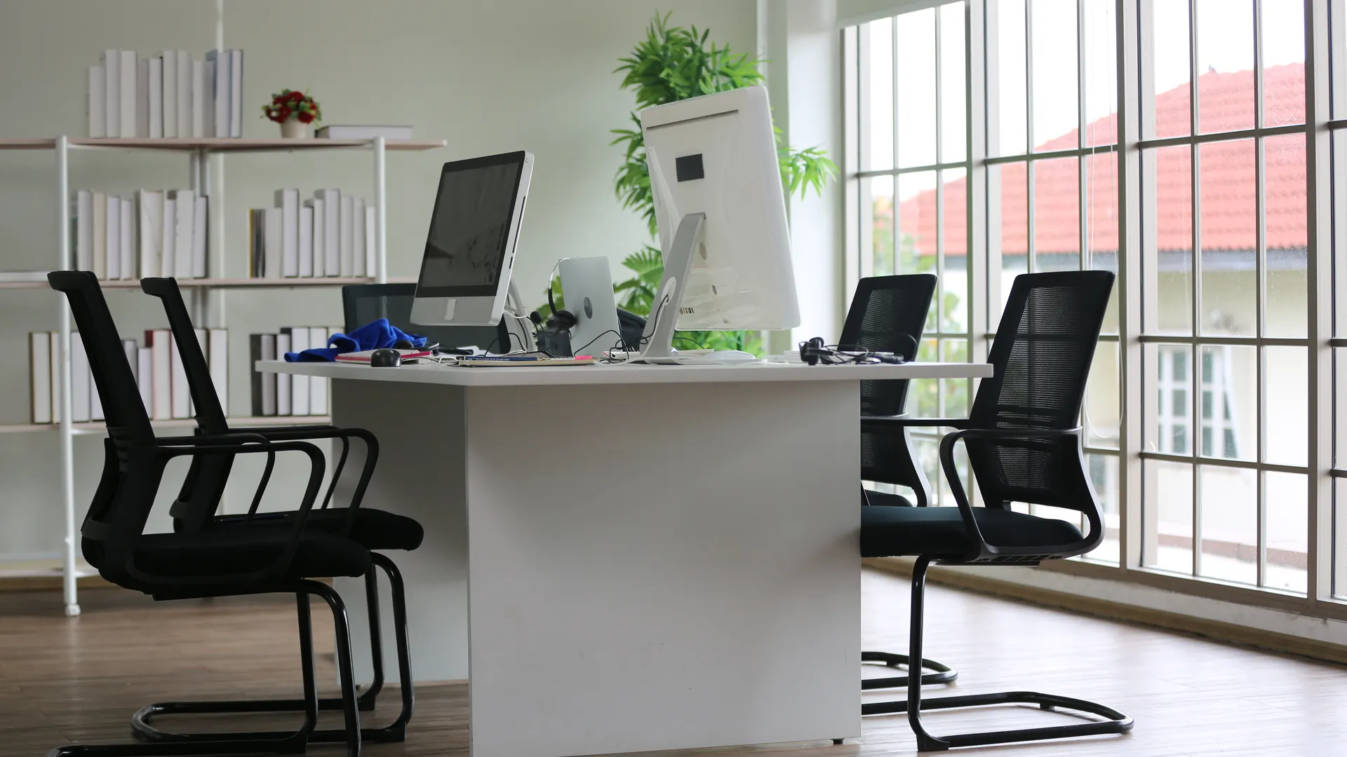 A bright open-plan office with a white desk, black mesh chairs, a computer and tall windows.