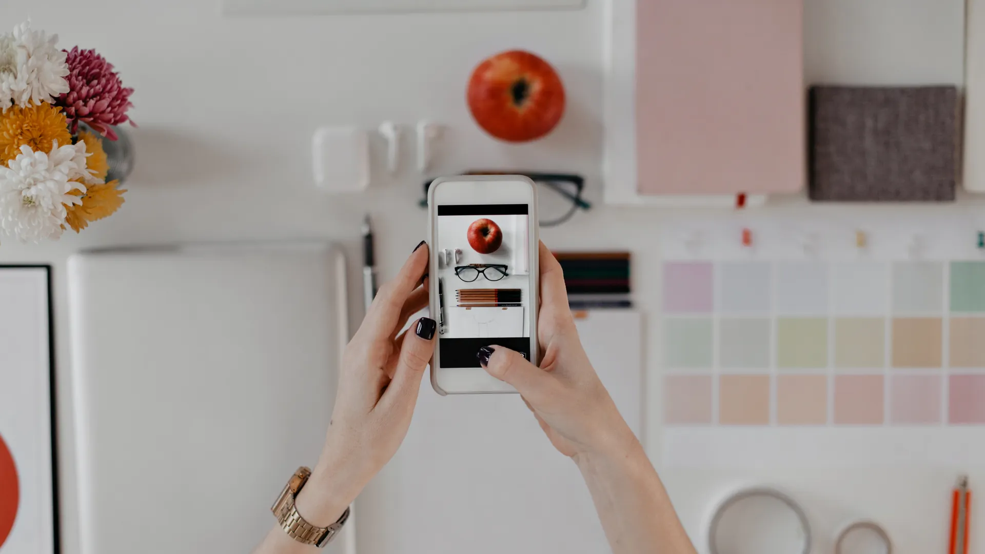 A pair of hands holds a smartphone over a styled desk of colour swatches, flowers and stationery to photograph a flat lay.