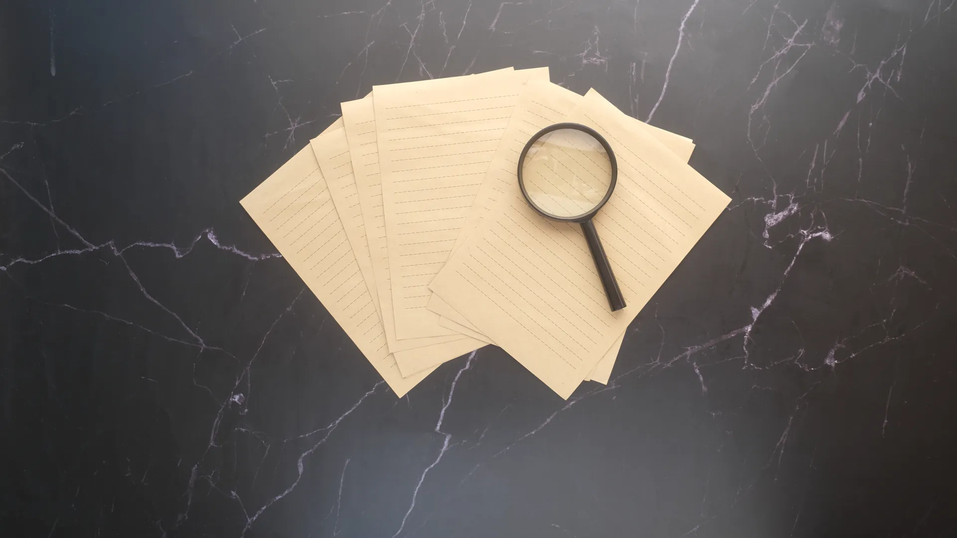 A magnifying glass rests on a pile of lined paper sheets on a dark marble surface.