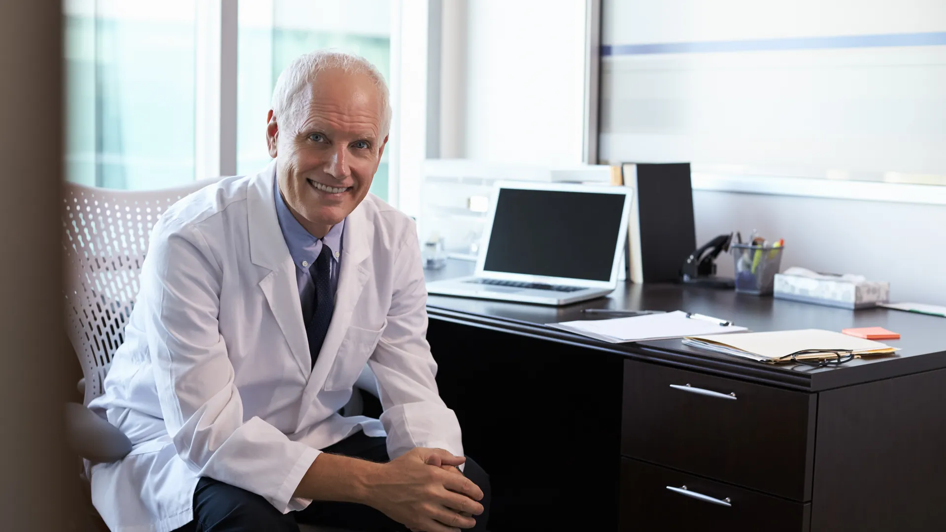 A grey-haired doctor in a white coat and tie sits smiling at a dark desk with a laptop and folders in a bright office.