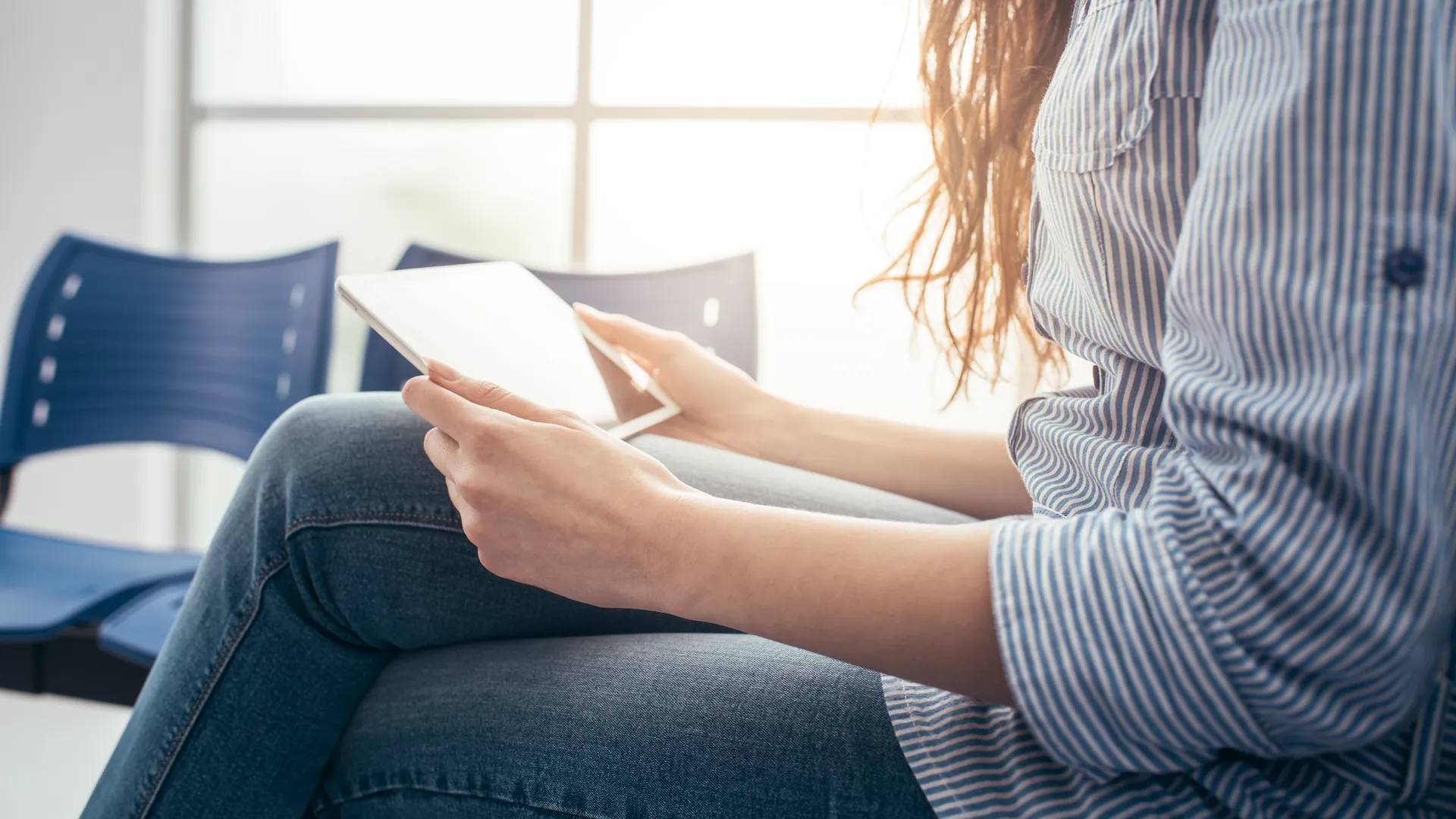 A person in a striped shirt and jeans holds a tablet on their lap while sitting on blue chairs in a sunlit waiting area.