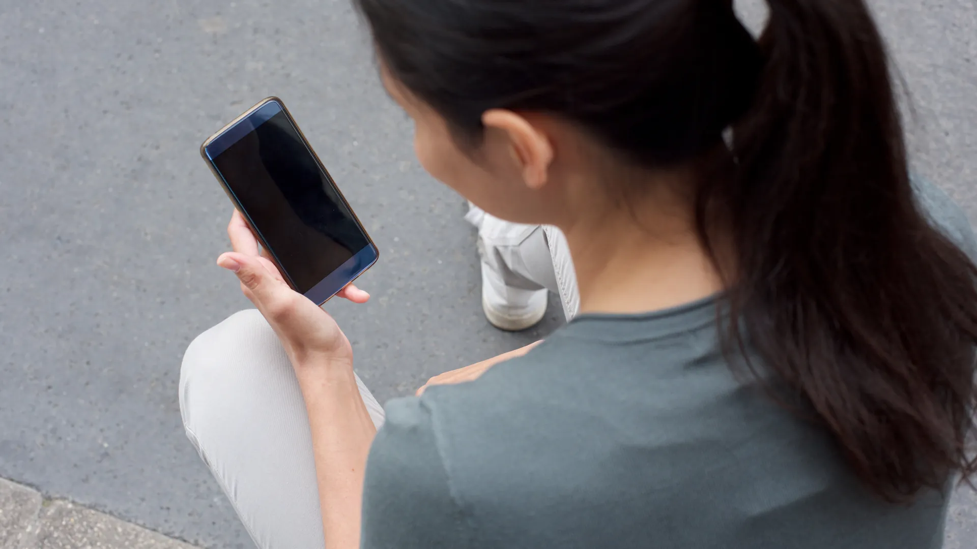A woman with a ponytail sits outdoors looking at a smartphone.