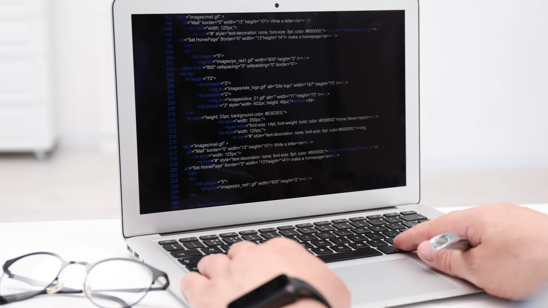 A person's hands typing on a laptop whose screen shows lines of HTML code, with glasses resting on the desk.