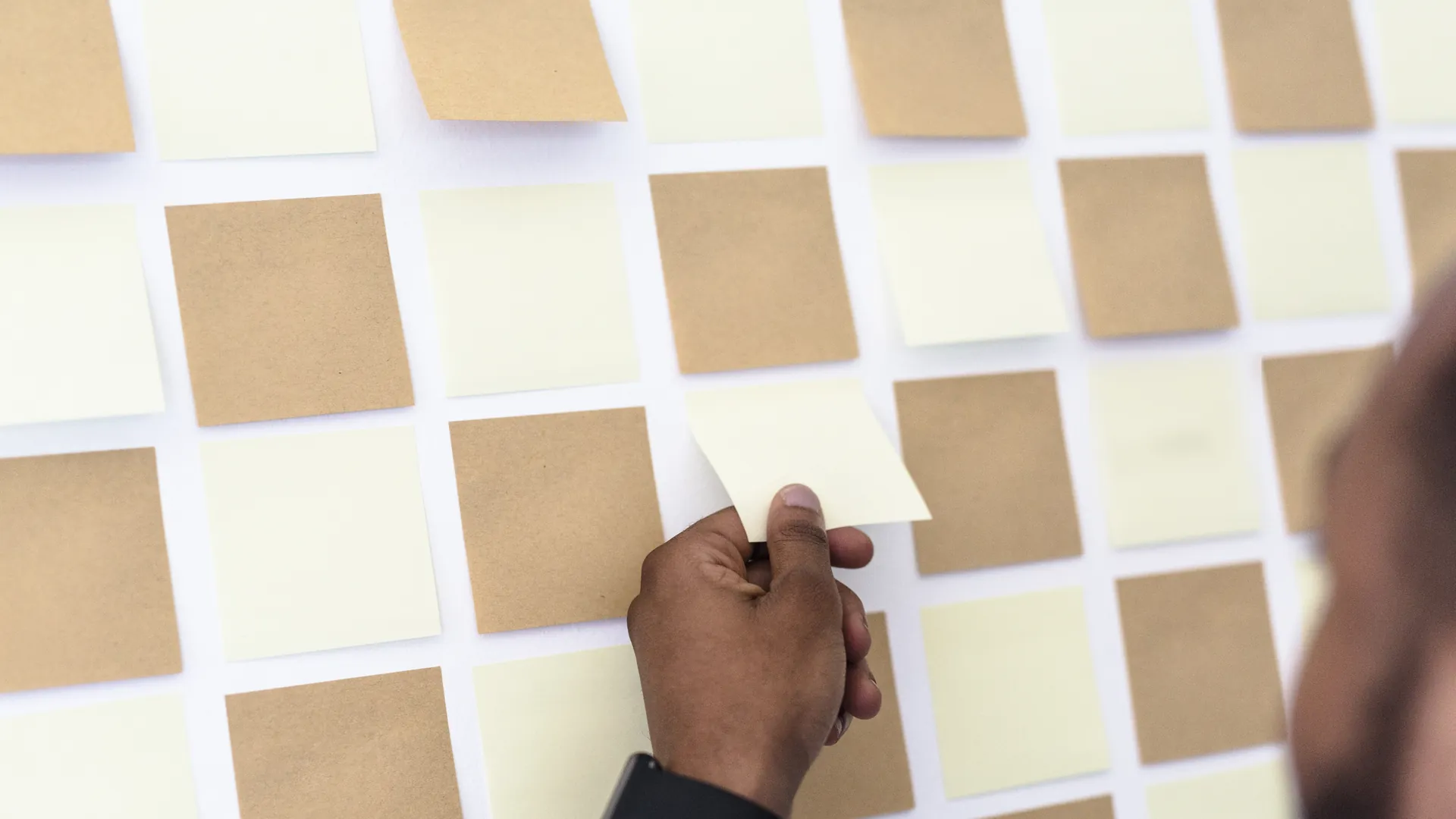 A hand peels a blank sticky note from a wall grid of blank yellow and brown notes.