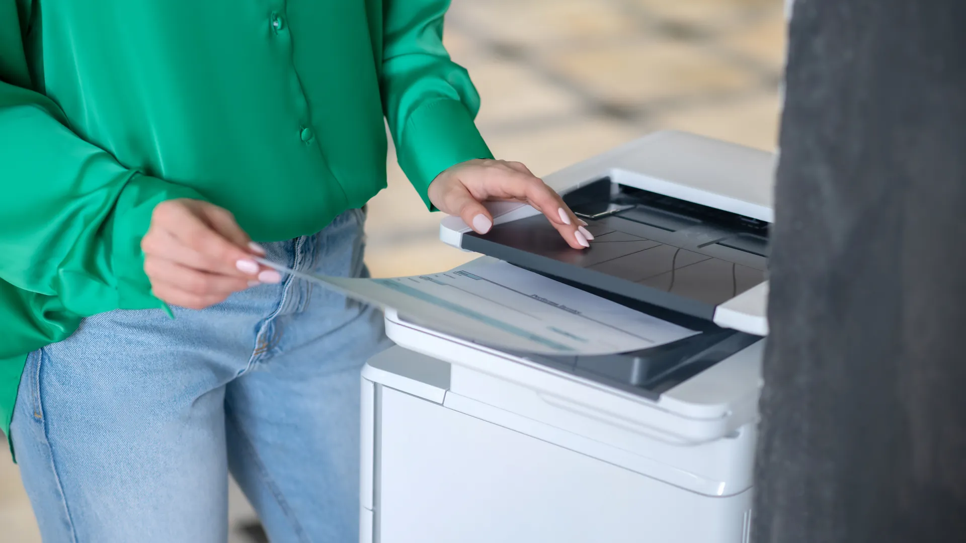 A woman in a green shirt and jeans places a sheet of paper on the glass of an office copier.