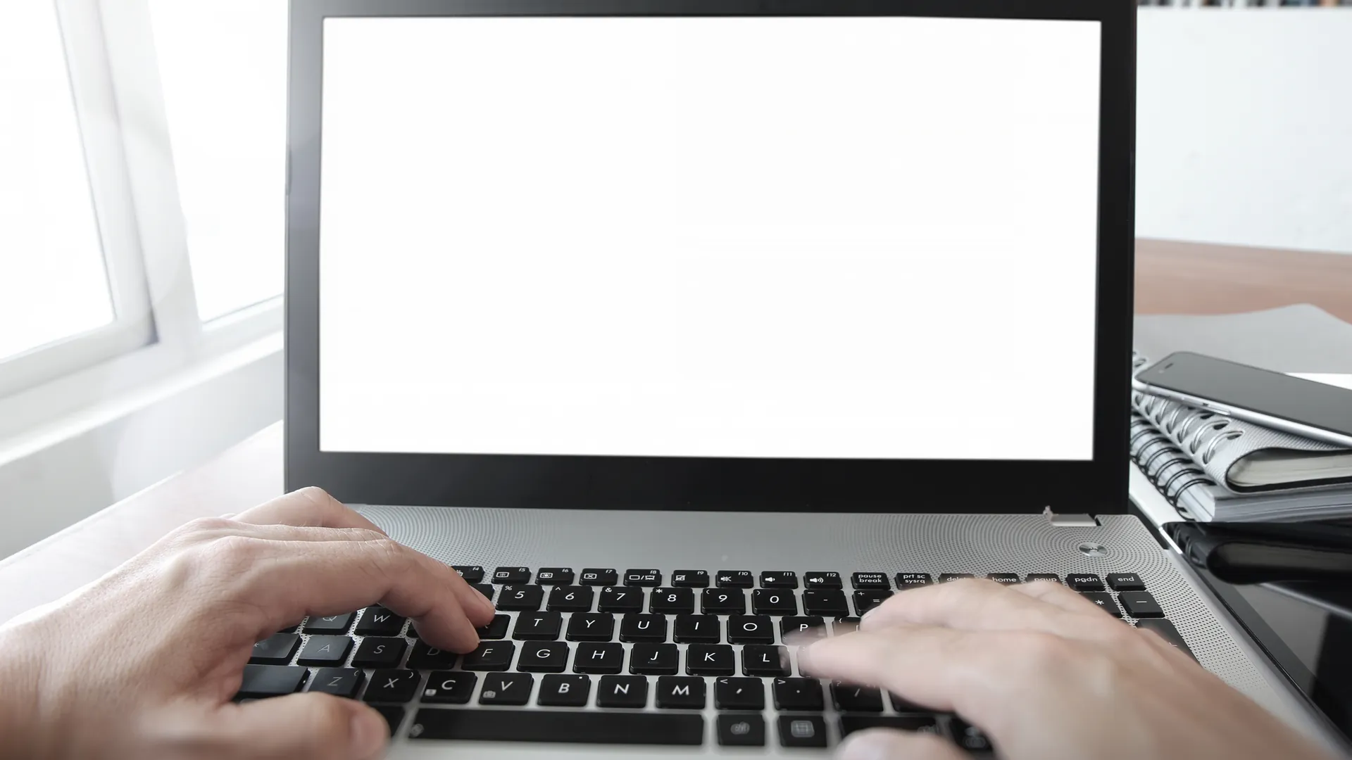 Two hands type on a laptop with a blank white screen beside a stack of notebooks on a desk.