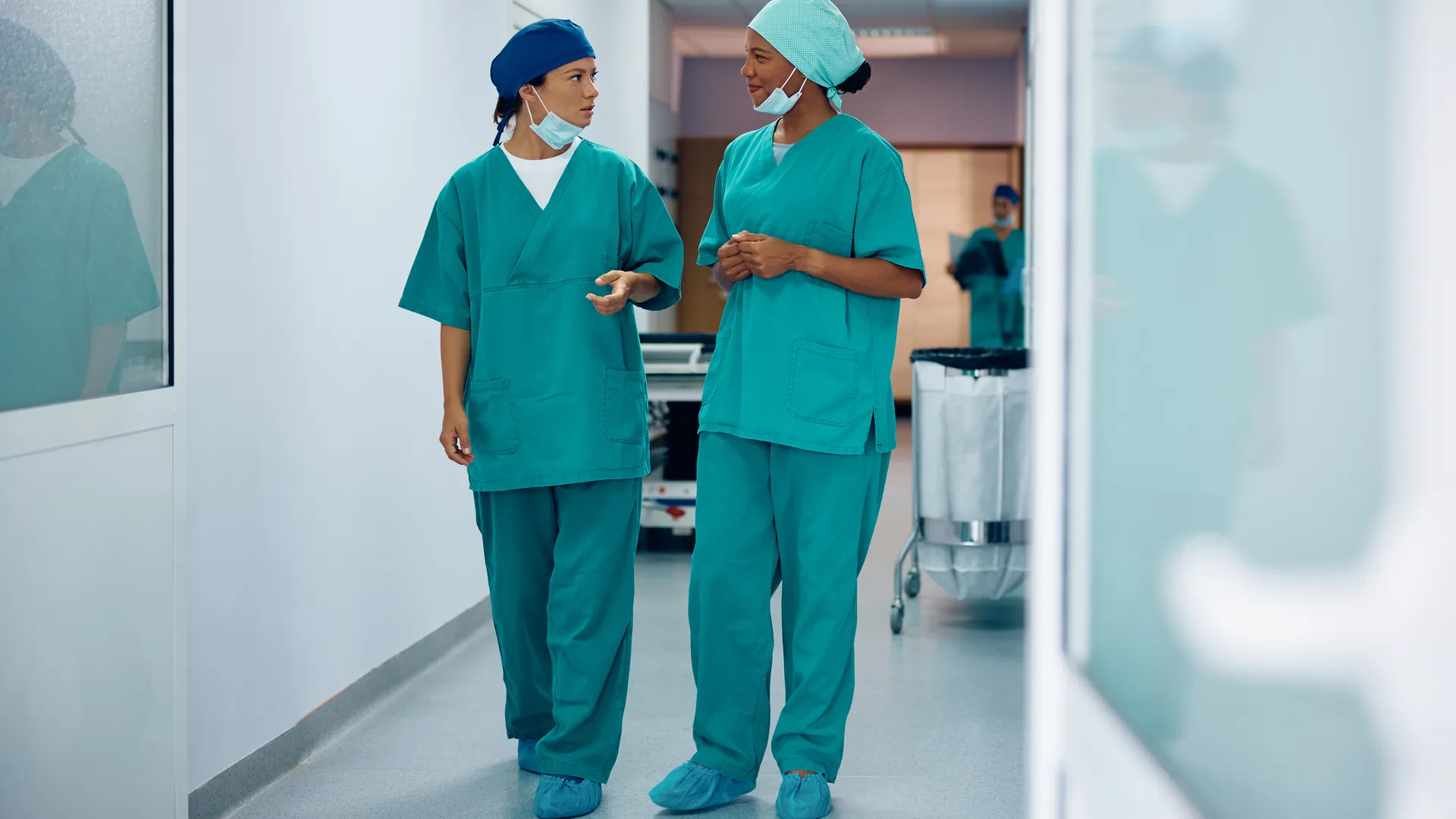 Two healthcare workers in green scrubs and surgical caps talk as they walk along a bright hospital corridor.
