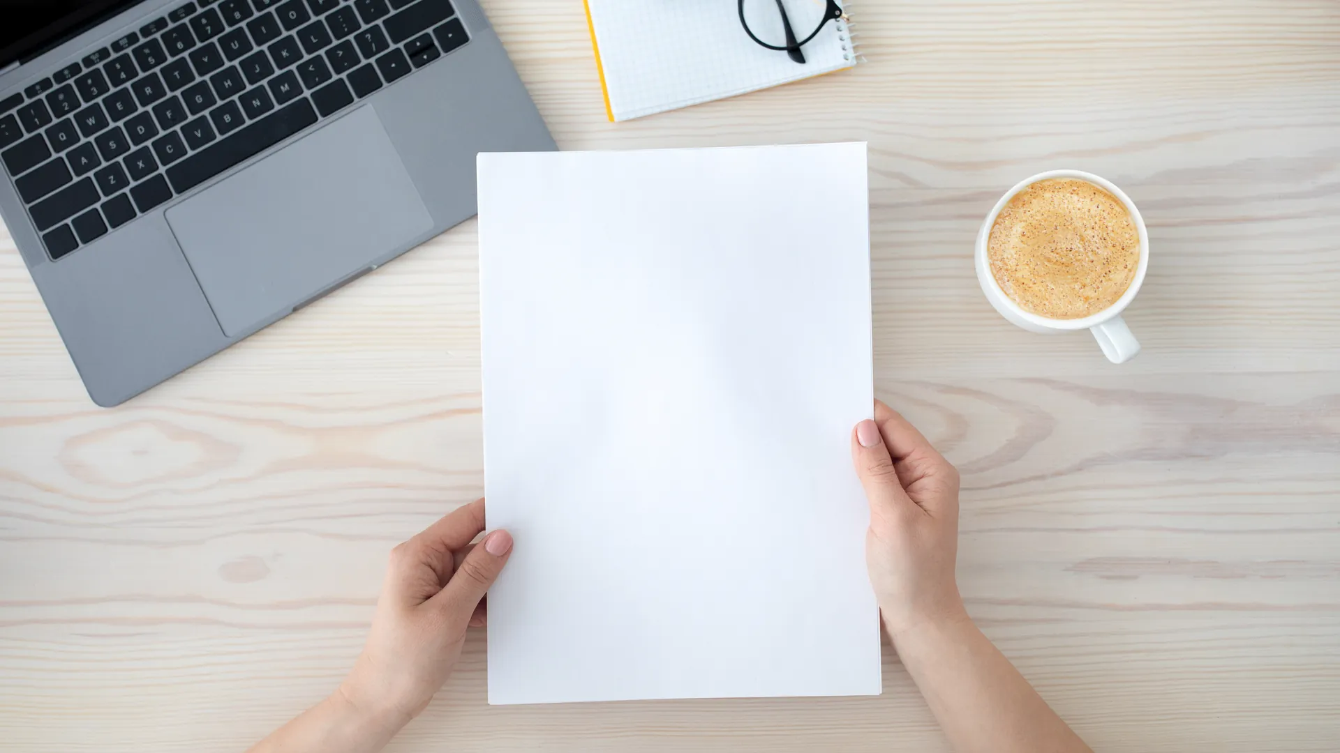A person holds a blank sheet of paper above a wooden desk with a laptop, a cup of coffee and a notebook.