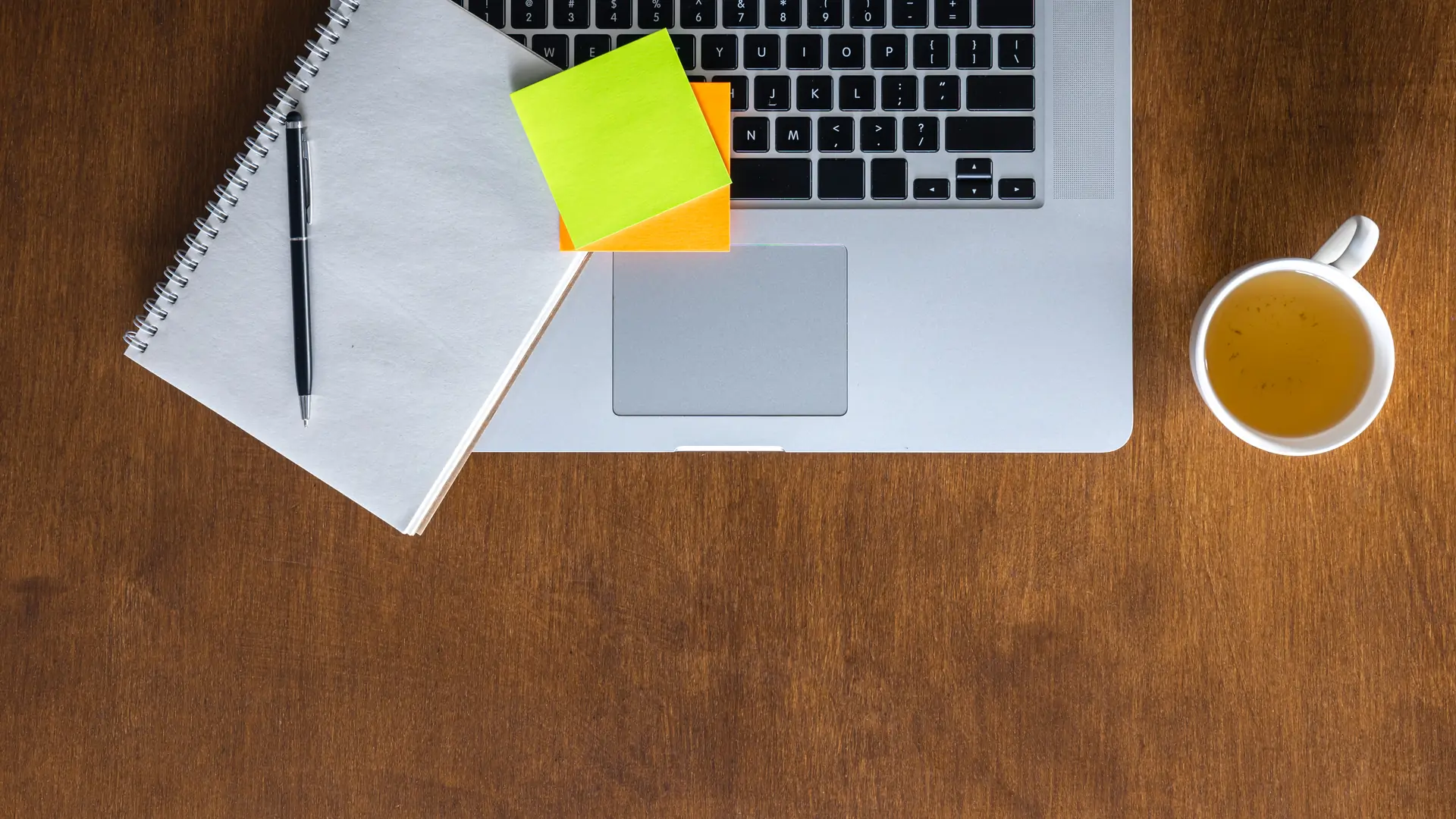 A top-down view of a laptop, notepad and pen, sticky notes and a cup of tea on a wooden desk.