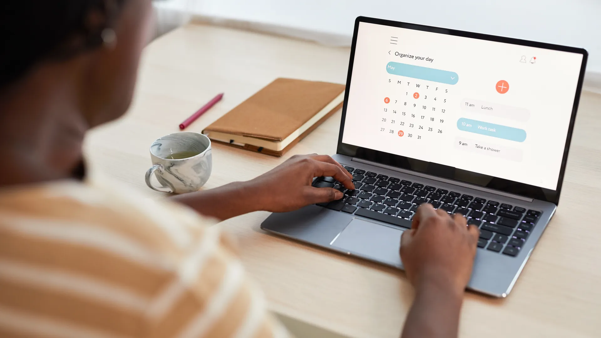 A woman working on a laptop that shows a digital calendar planner, with a cup and notebook beside her.