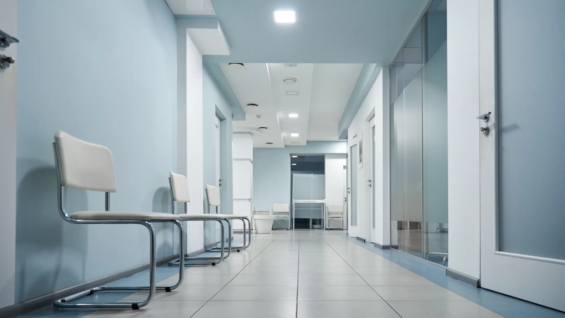 An empty hospital corridor with pale blue walls and a row of white chairs along one side.