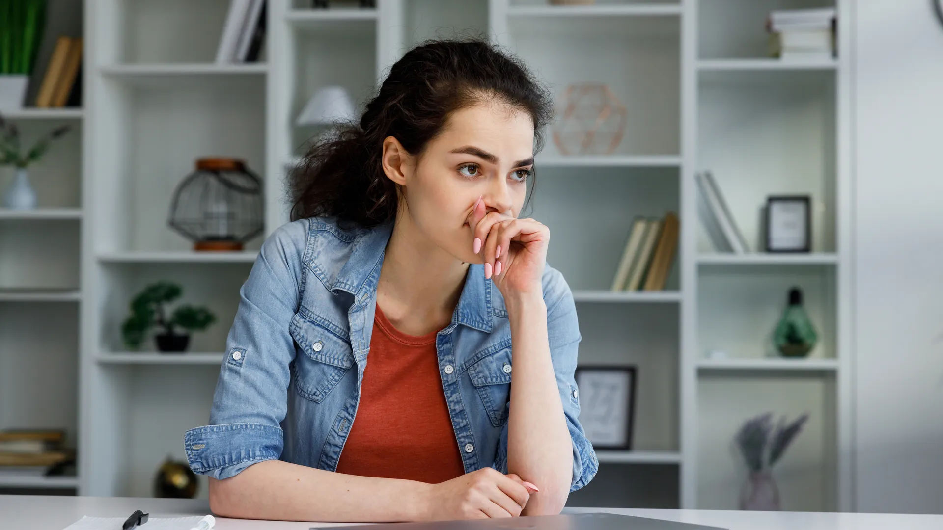 A young woman in a denim jacket rests her hand against her face, looking worried, at a table in front of white shelves.